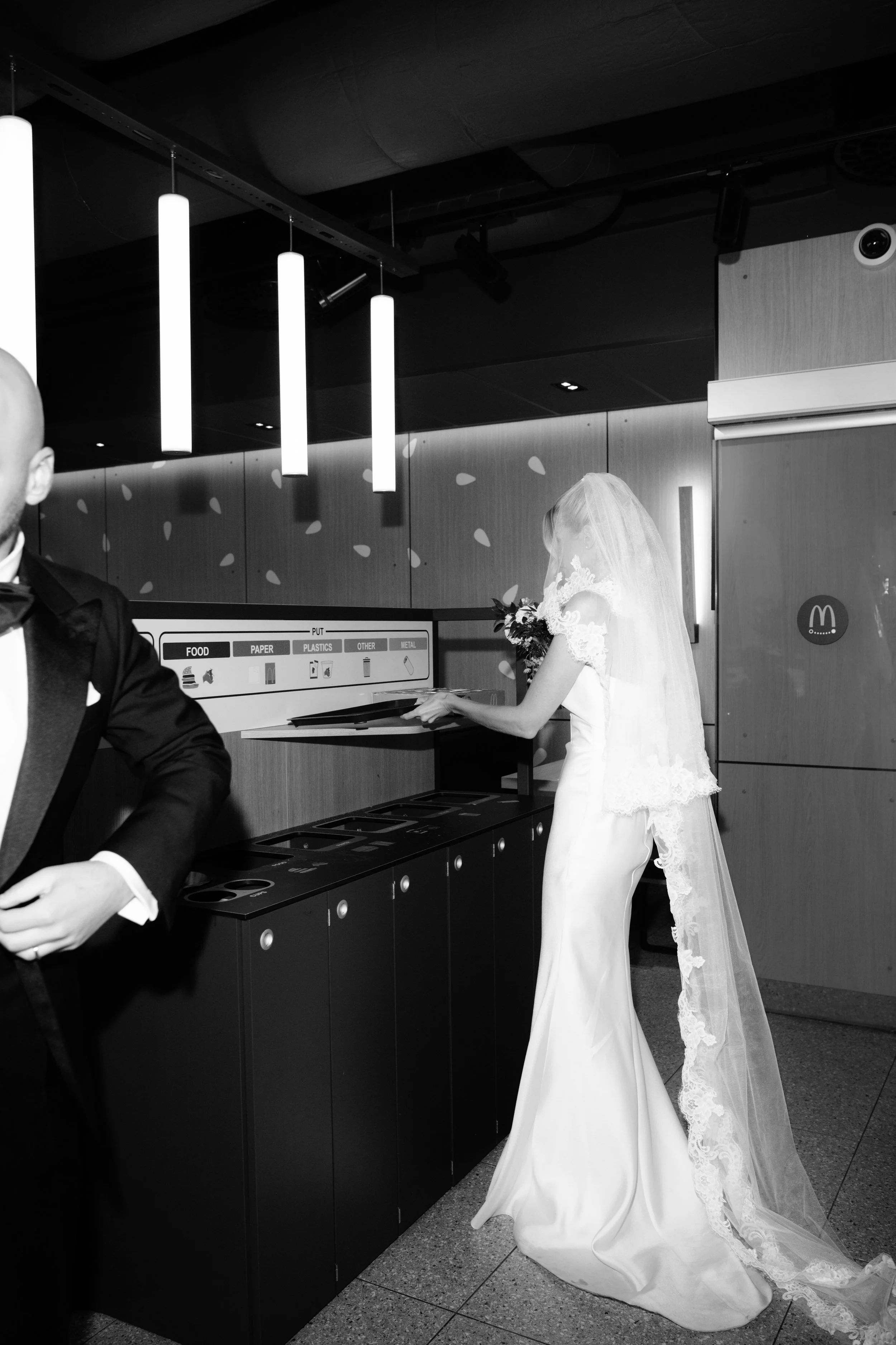Black and white photo of a bride in a wedding dress and veil, placing a tray in a recycling bin at a McDonald's restaurant, with a man in a tuxedo partially visible in the foreground.