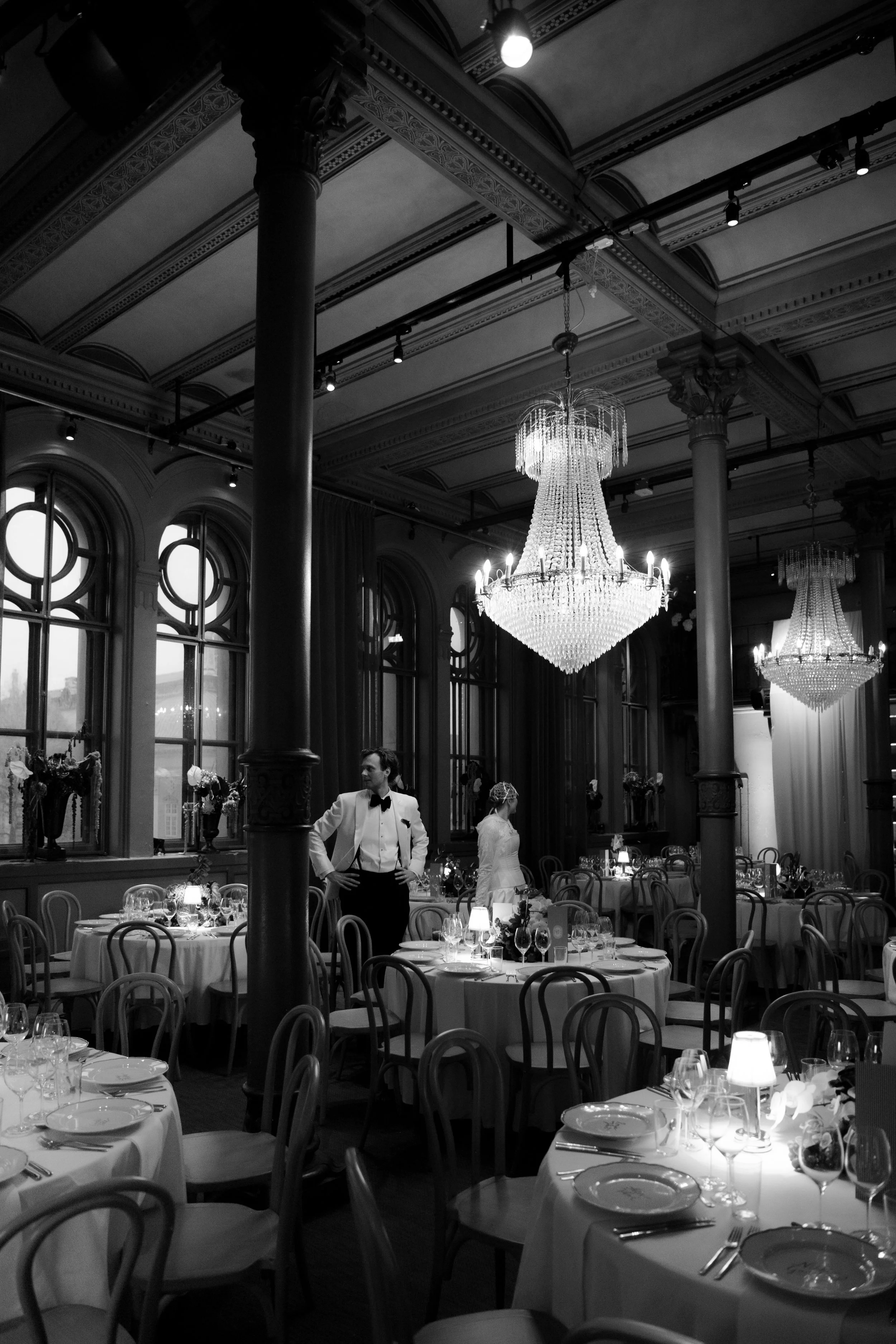 Black and white image of an elegant, upscale banquet hall with large chandeliers, tall arched windows, and tables set for a formal event. There are two waitstaff, a man in a tuxedo and a woman in a white dress, preparing the tables.