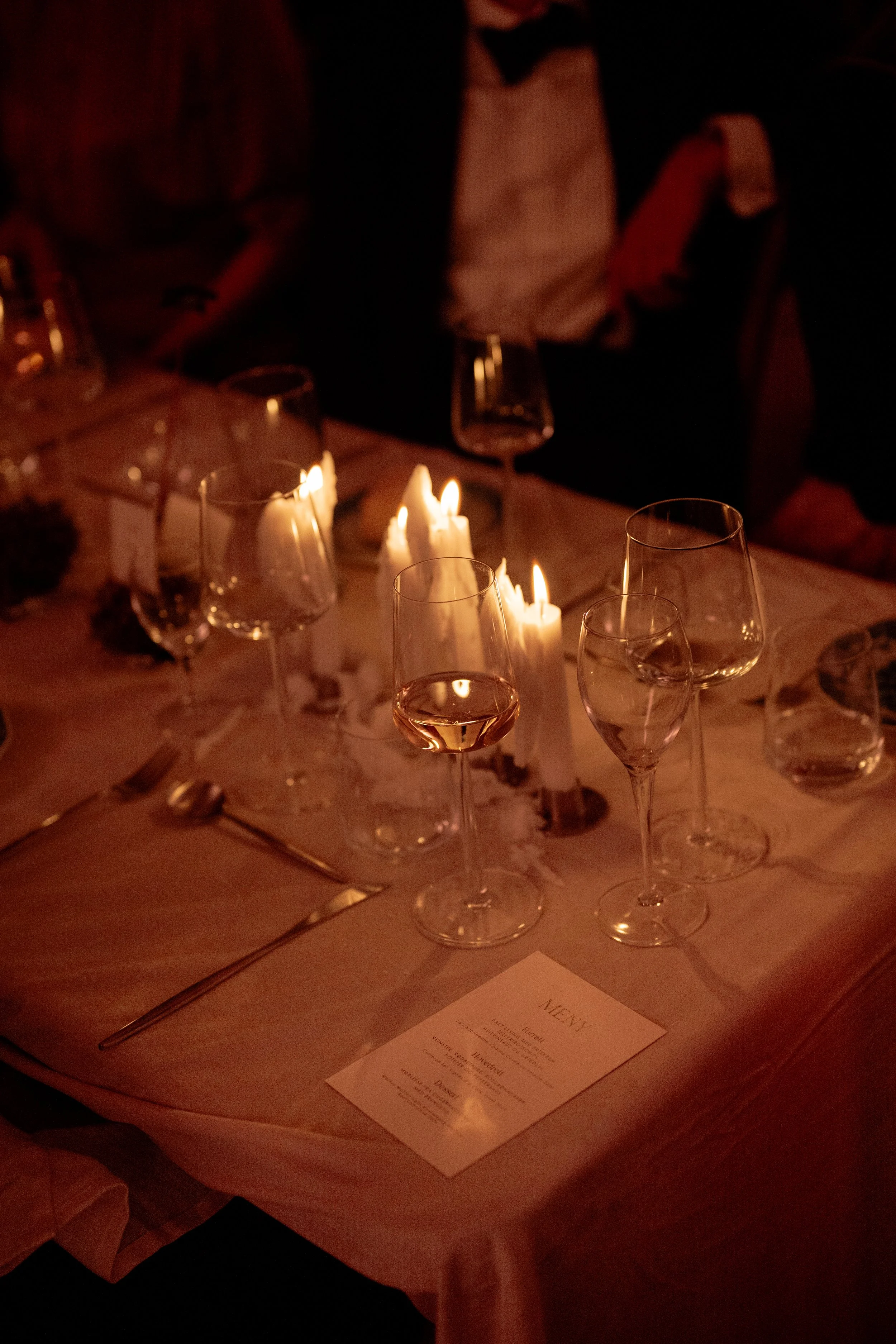 A dimly lit candlelit dinner table with empty wine glasses, a menu, and utensils. There are people in formal attire seated around the table.