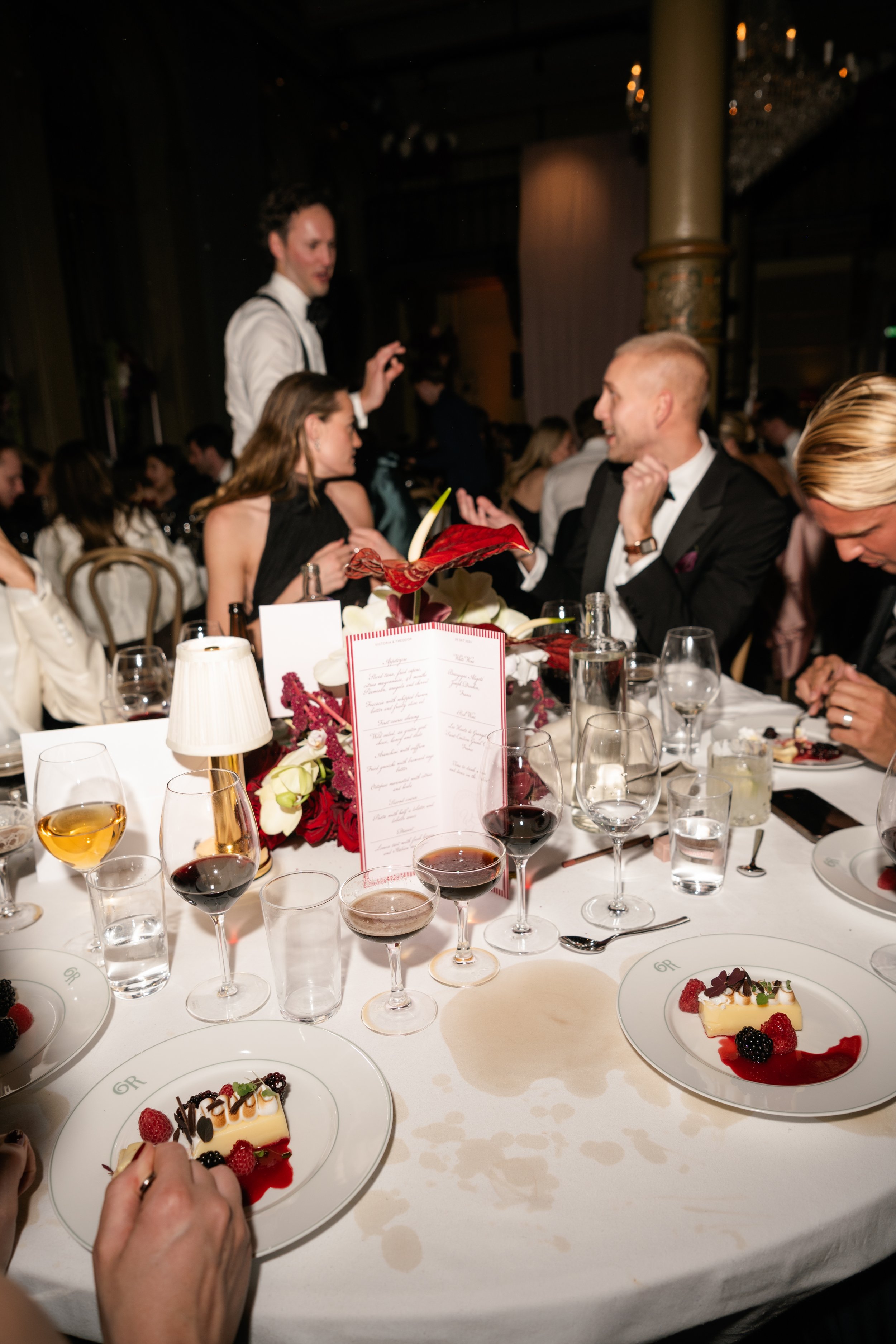 A formal dinner party with guests in tuxedos and evening dresses seated around a table with various desserts and drinks. A man in a white shirt and black suspenders is standing and speaking to a woman in a black dress. The table is decorated with flo