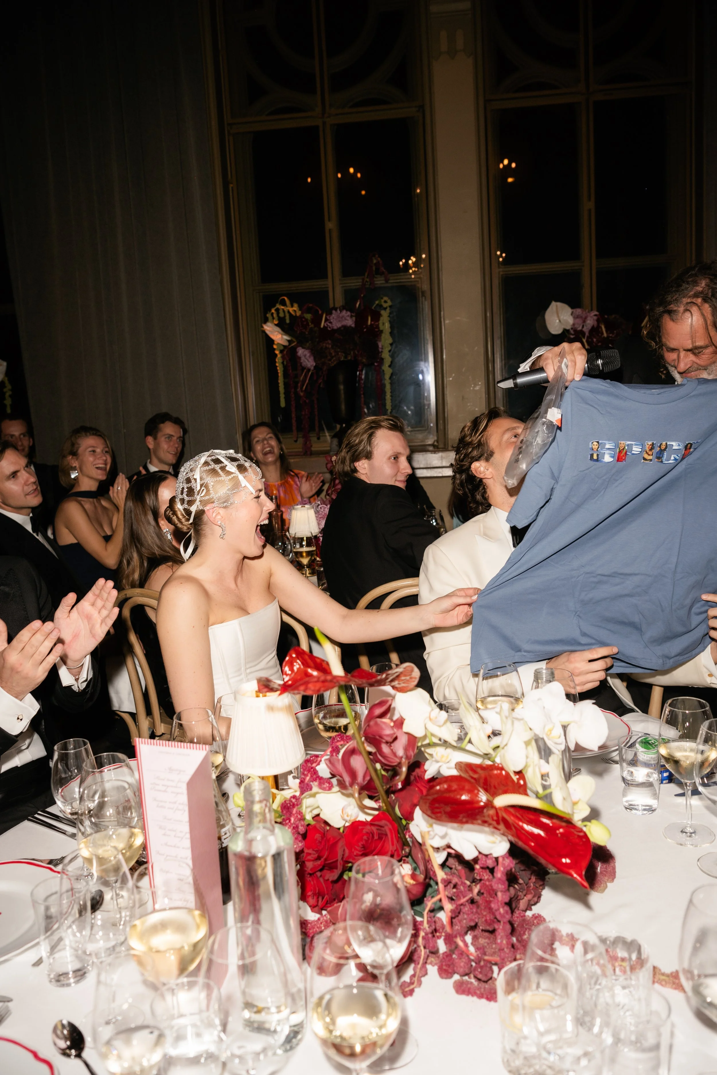 Guests at a wedding reception laughing and celebrating, with a woman in a white dress and a lace headpiece in the foreground.