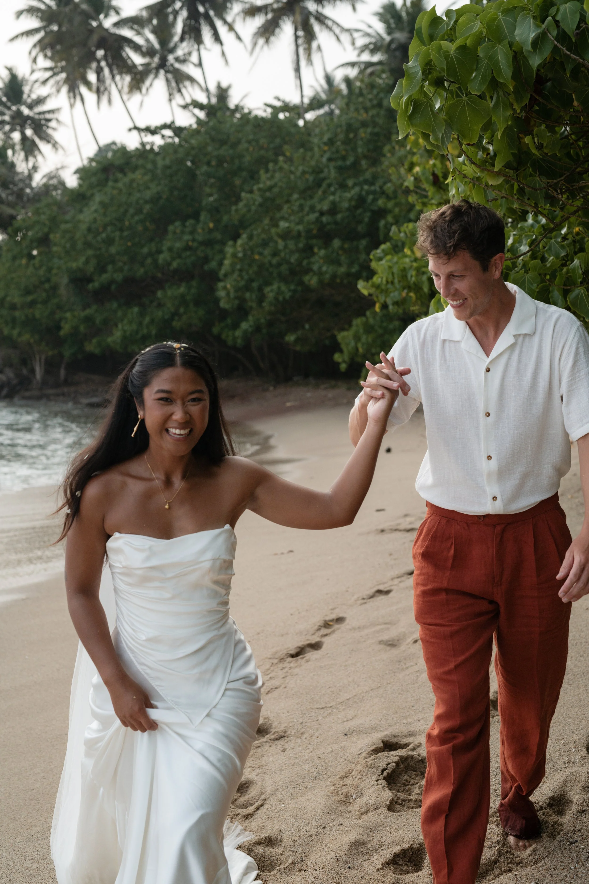 A smiling woman in a white wedding dress and a man in a white shirt and rust-colored pants dancing on a sandy beach surrounded by lush green trees.