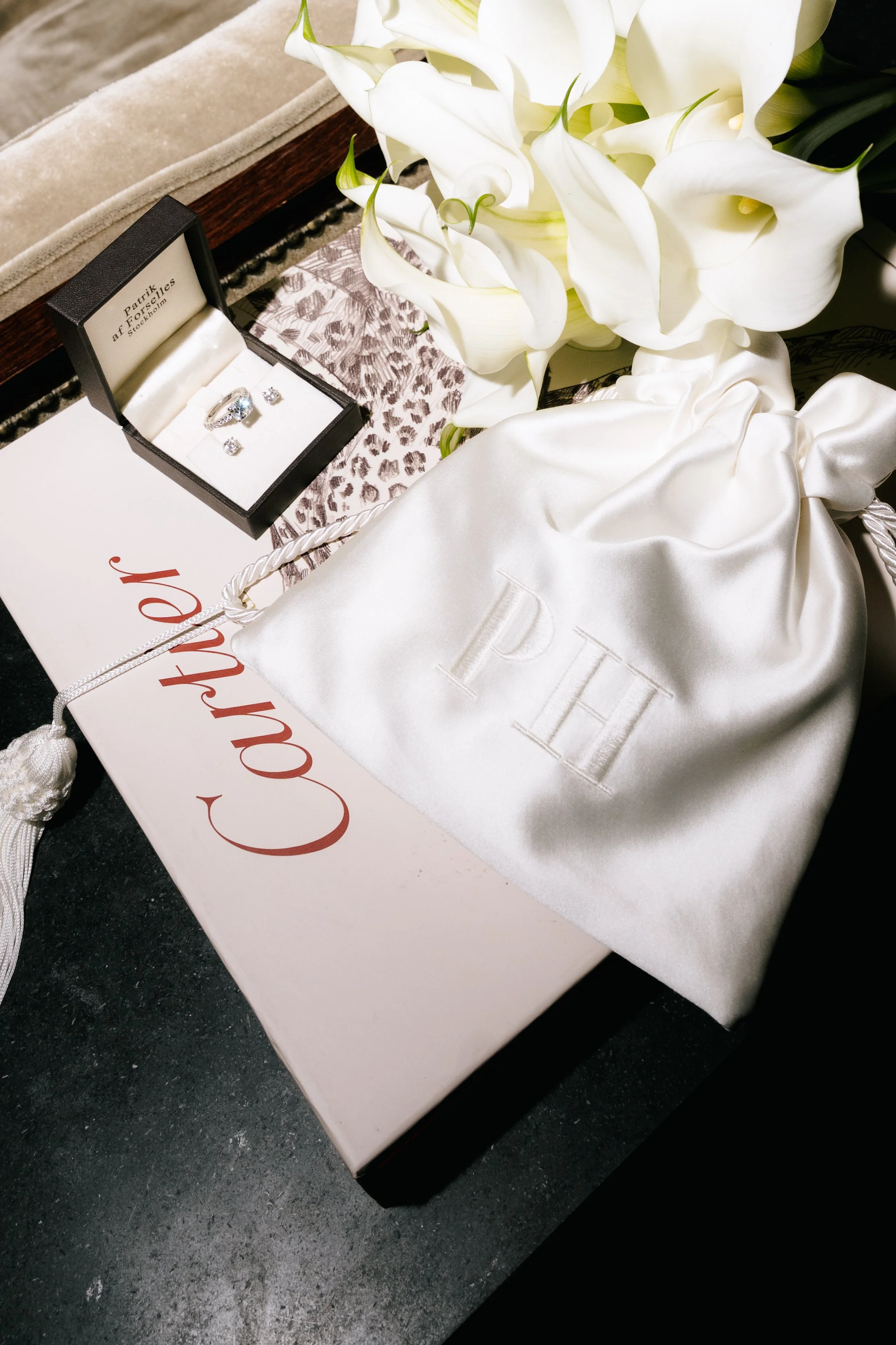 A white Cartier gift box, a white satin drawstring bag with the Cartier logo, a bouquet of white calla lilies, and a small black jewelry box with diamonds inside.