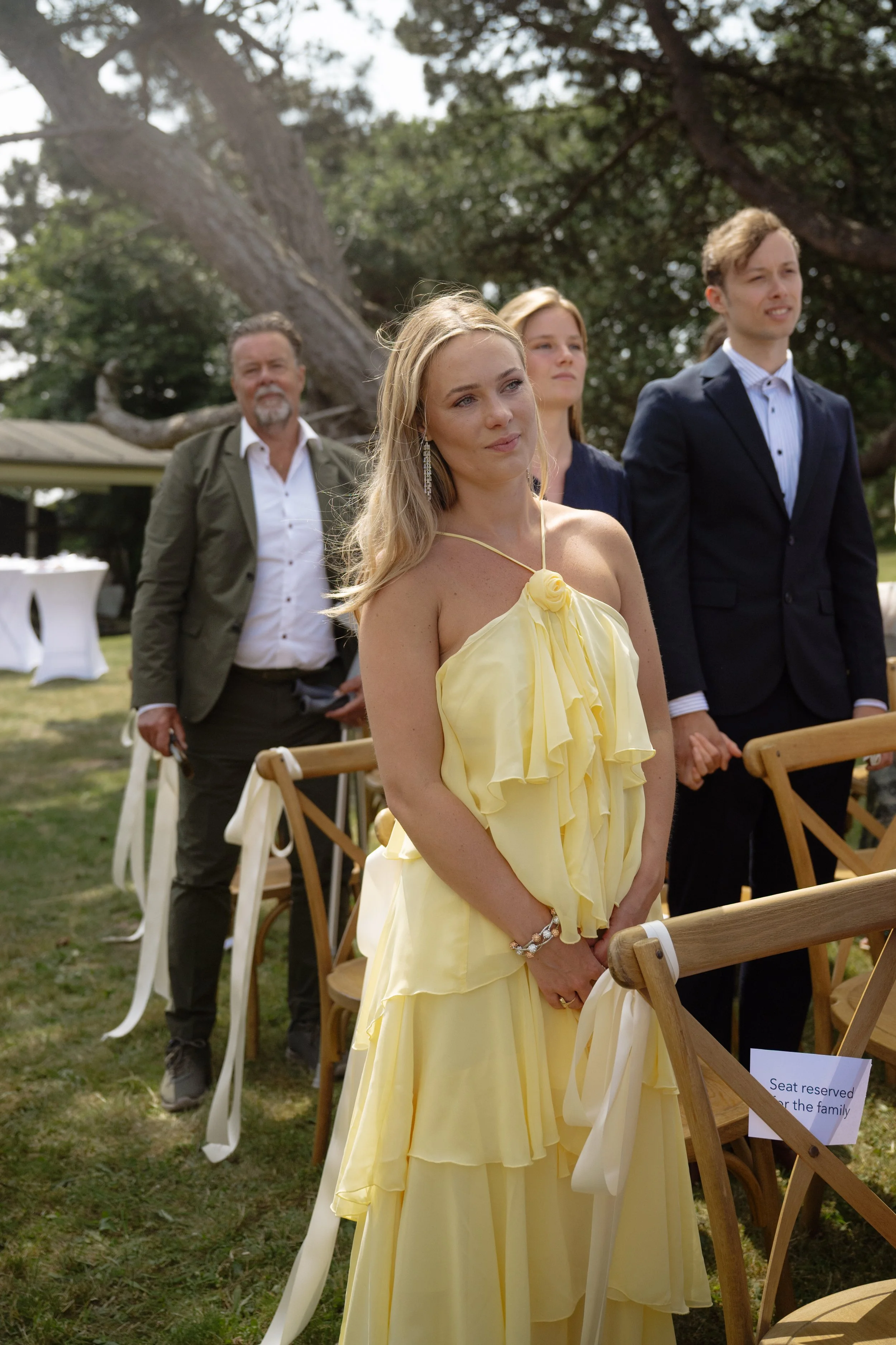 People attending a wedding ceremony outdoors, with a woman in a yellow dress standing in the foreground and others in formal attire in the background, under large trees.