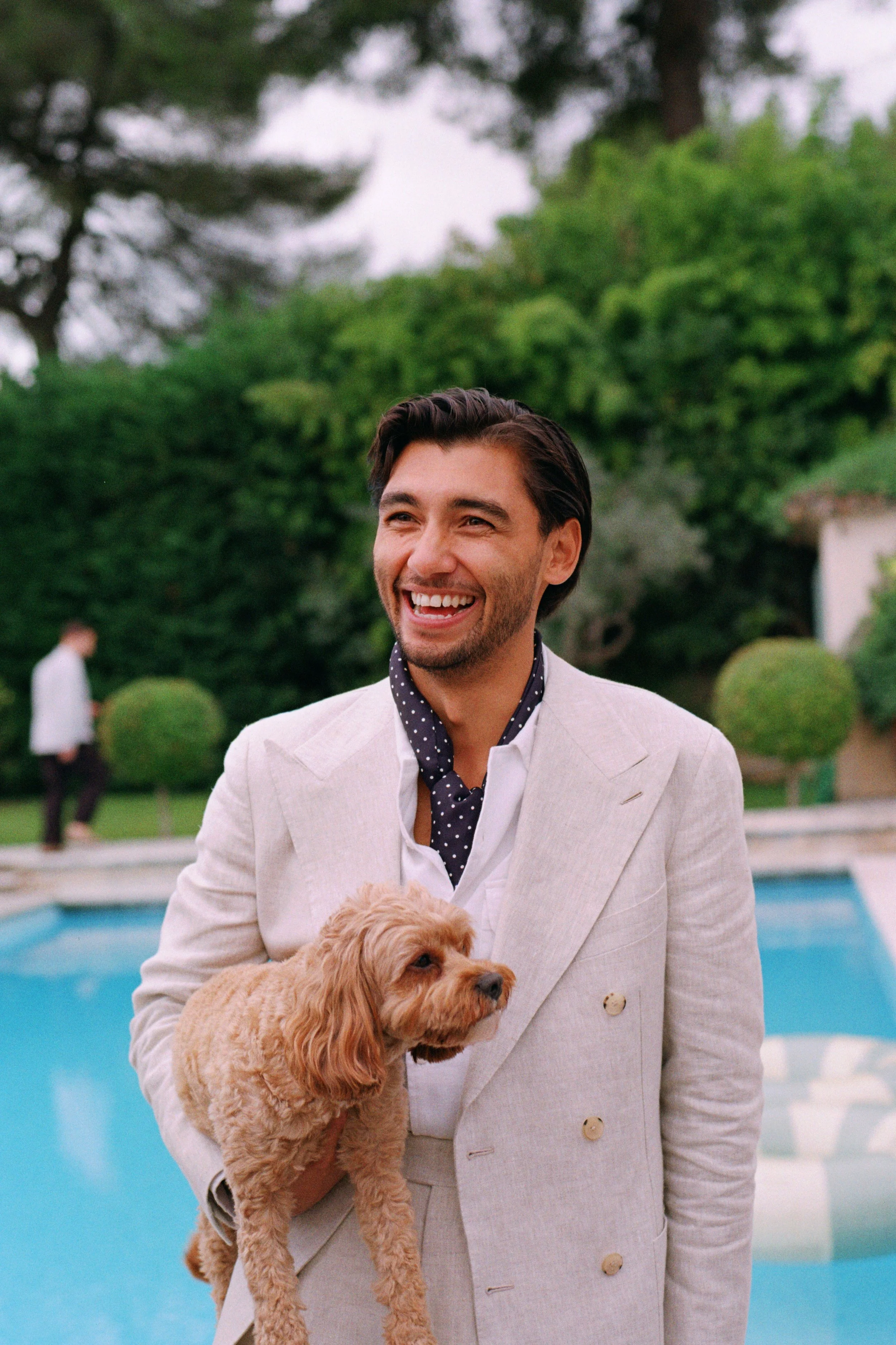 A man in a light-colored suit holding a small curly-haired dog, smiling near a swimming pool with greenery in the background.