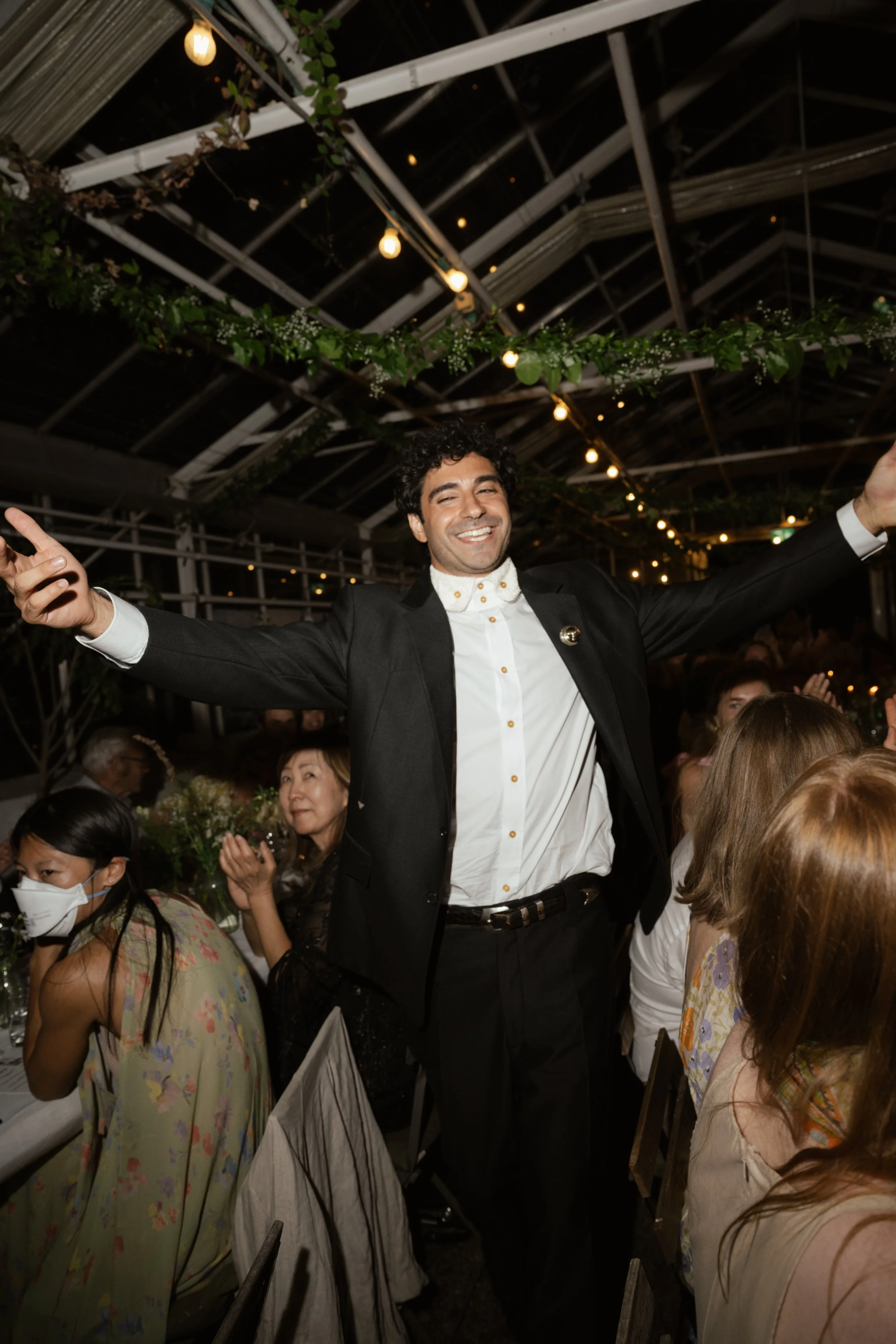 A man at a celebration or party with arms spread, smiling, surrounded by seated guests in an indoor venue decorated with string lights and greenery.