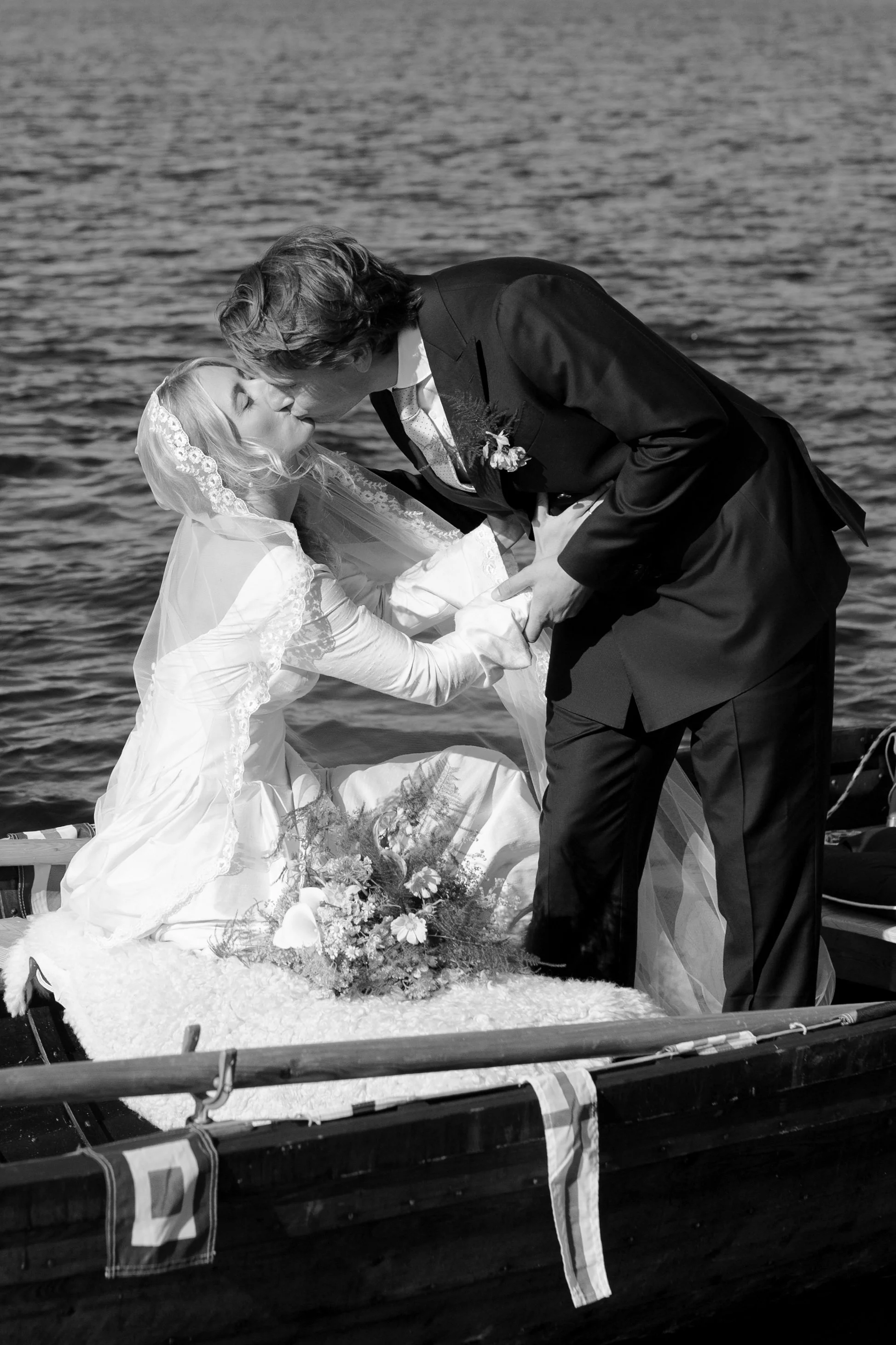 A bride and groom sharing a kiss on a boat by the water, with the bride sitting and the groom leaning over to kiss her.