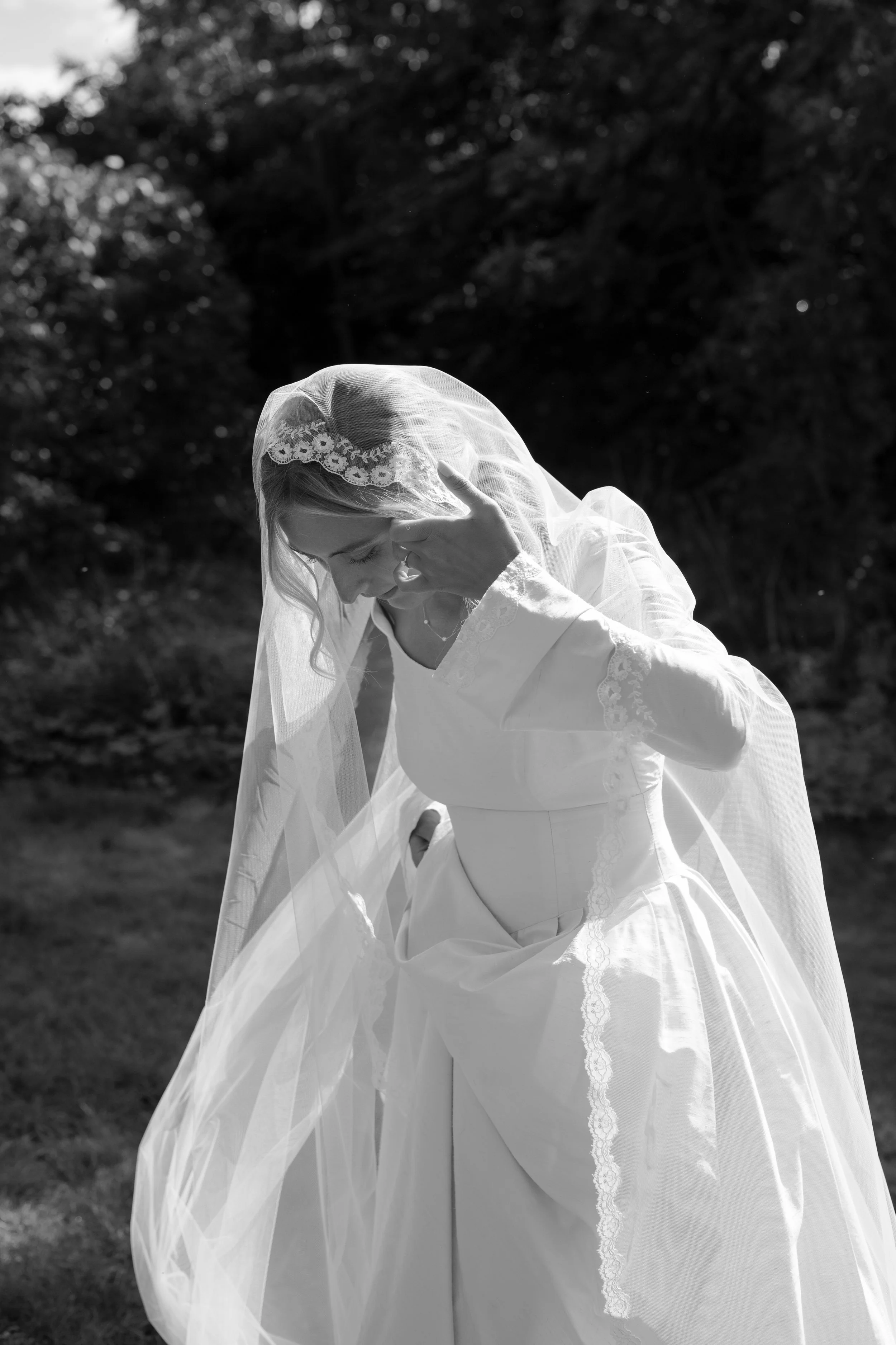 Black and white photo of a bride in a wedding dress with a long veil outdoors, holding her face with one hand and smiling.