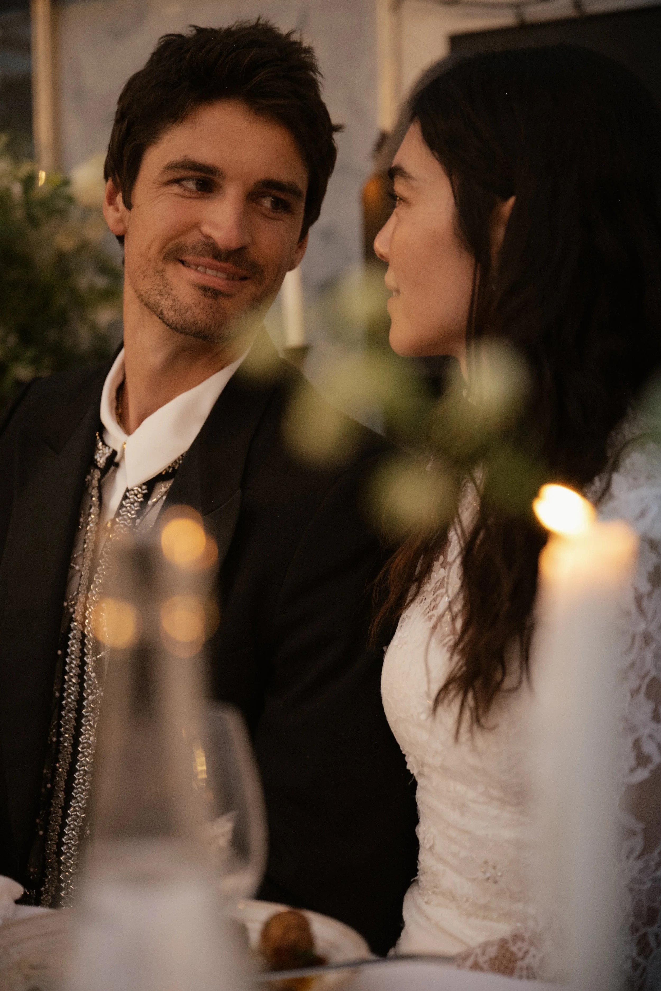 A man in a tuxedo looks at a woman in a white lace dress during a dinner event, with candles and blurred decorations in the background.