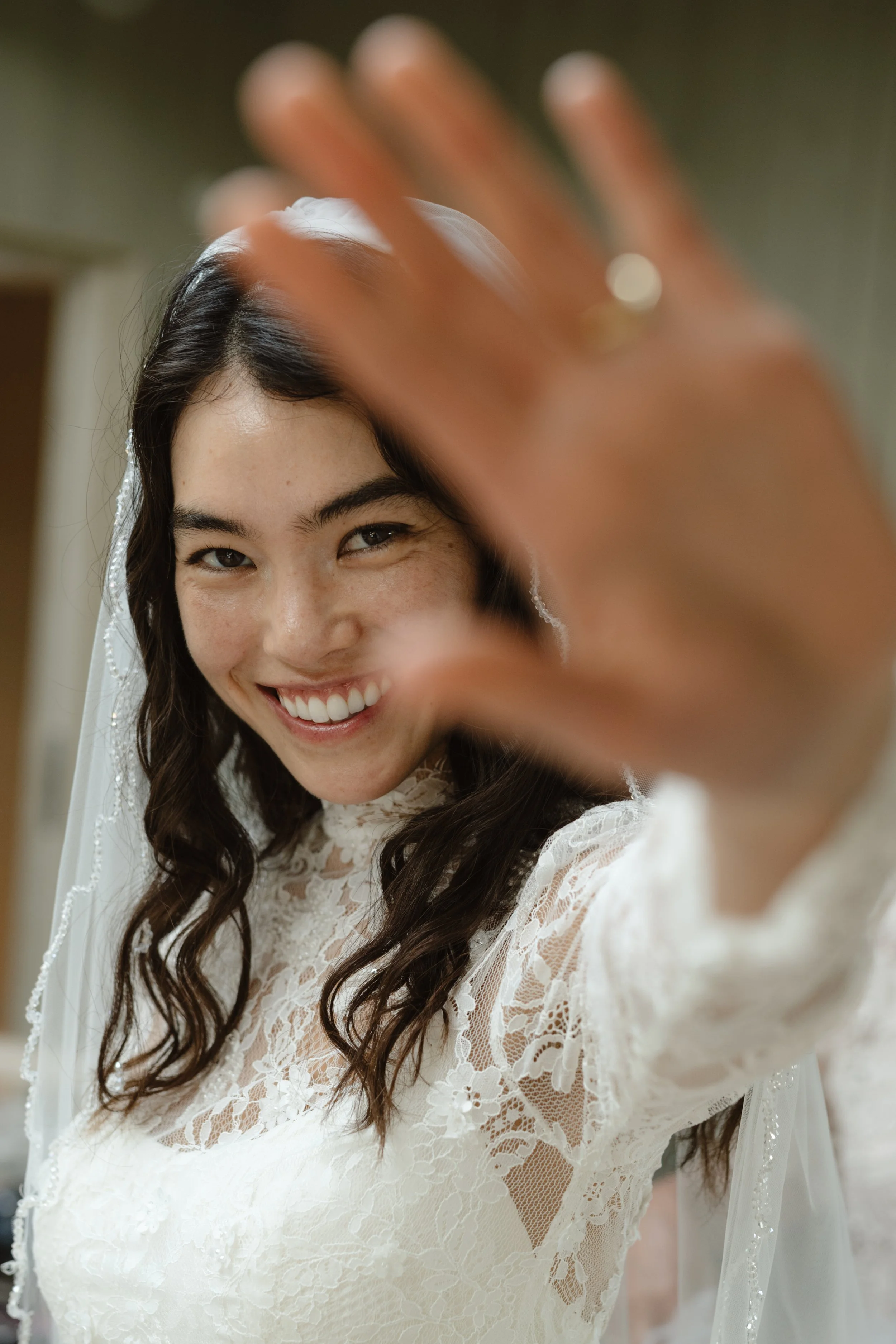 A young woman with dark wavy hair, wearing a white lace dress and a veil, smiling and reaching out towards the camera with her hand.