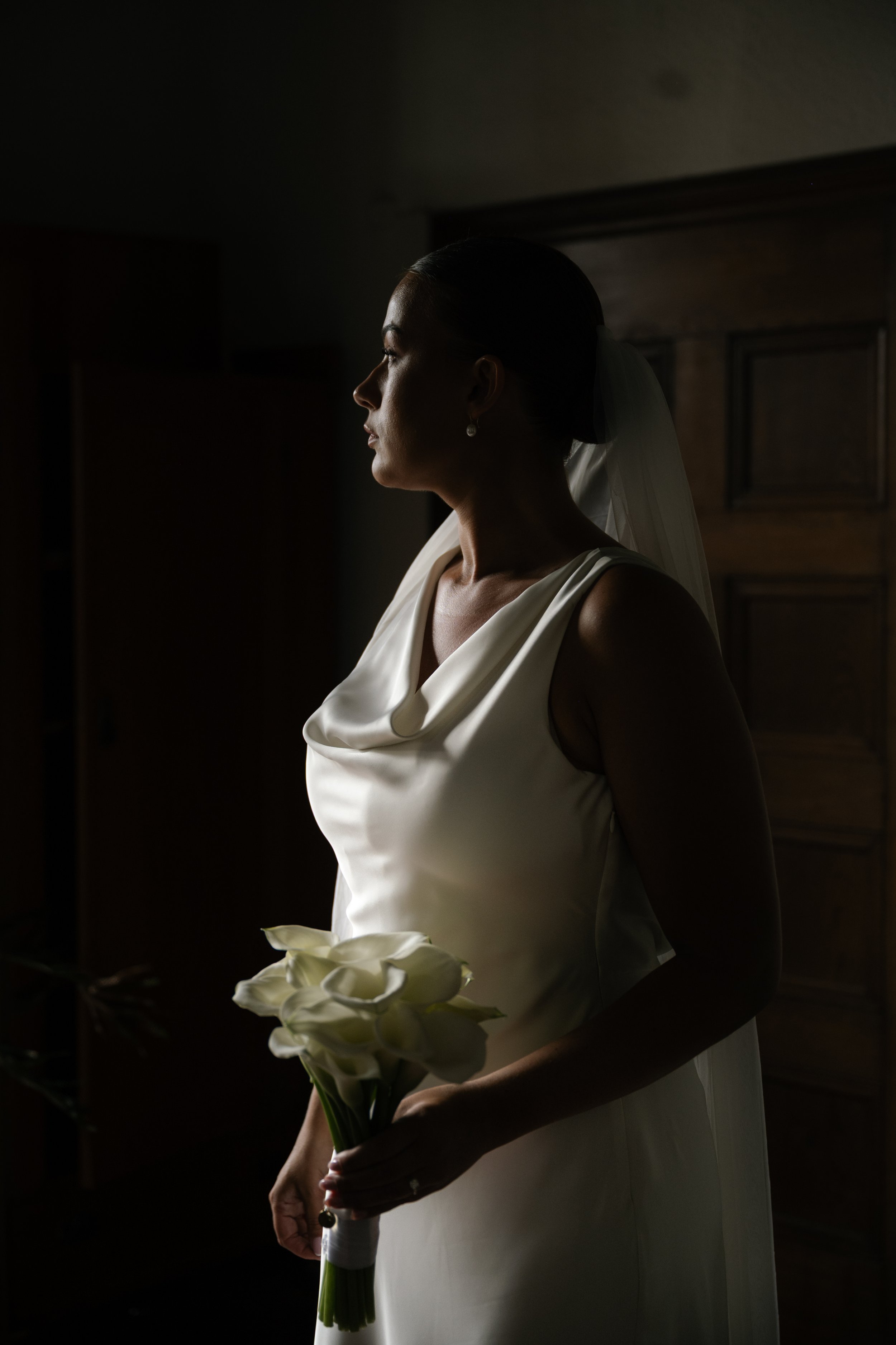 A bride in a white satin dress holding a bouquet of calla lilies, standing in a dimly lit room with wooden furniture, looking to the side.