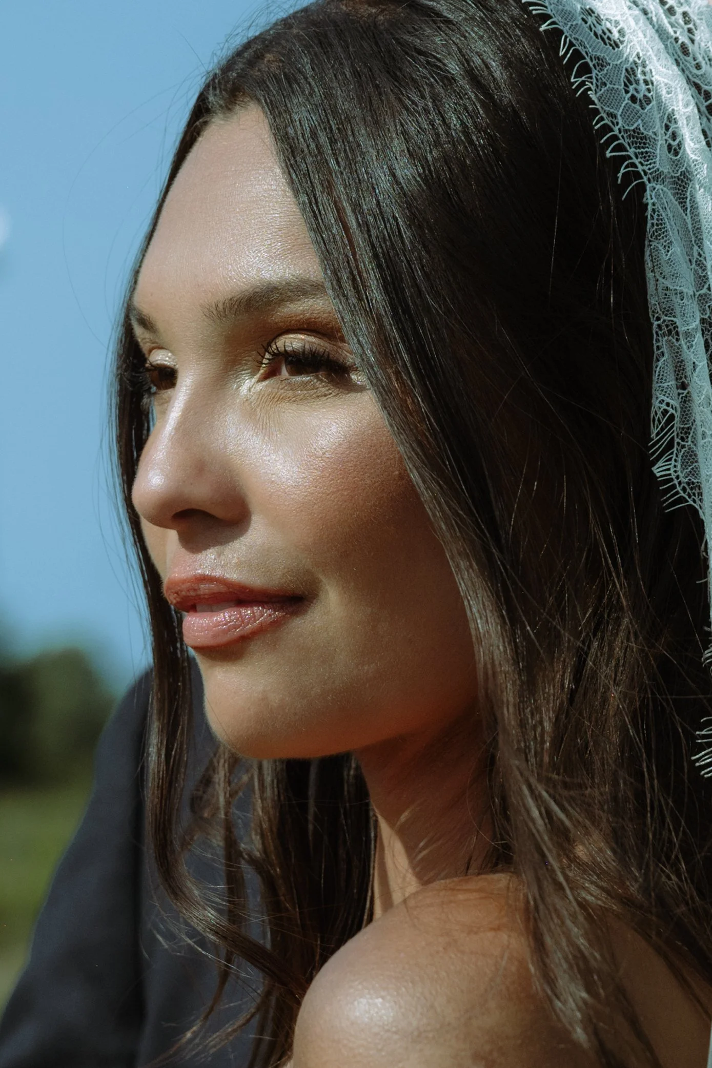 Close-up of a young woman with long, dark hair, wearing a white lace veil, outdoors on a sunny day.
