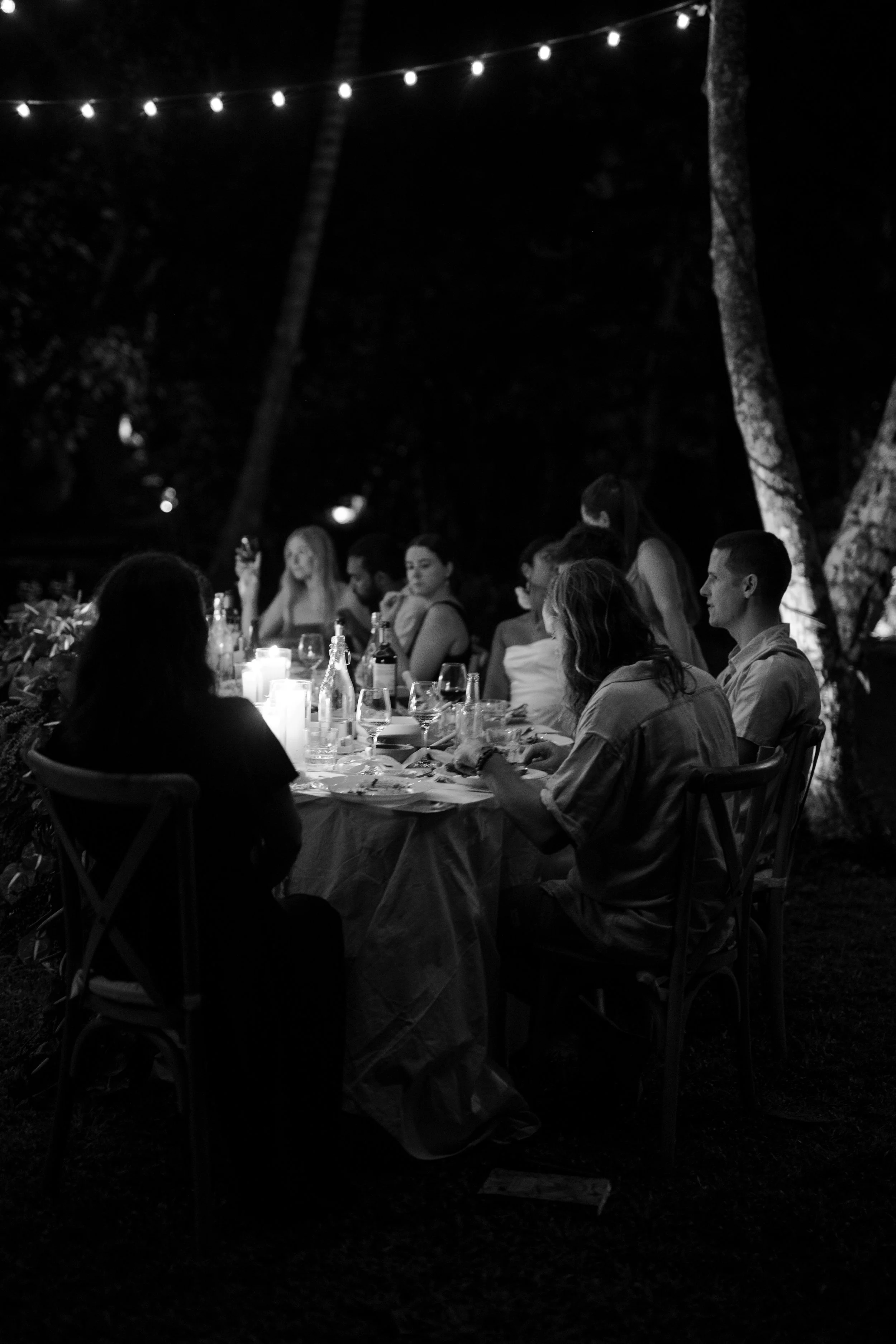 A group of people sitting around a dinner table outdoors at night, illuminated by candles and overhead string lights.