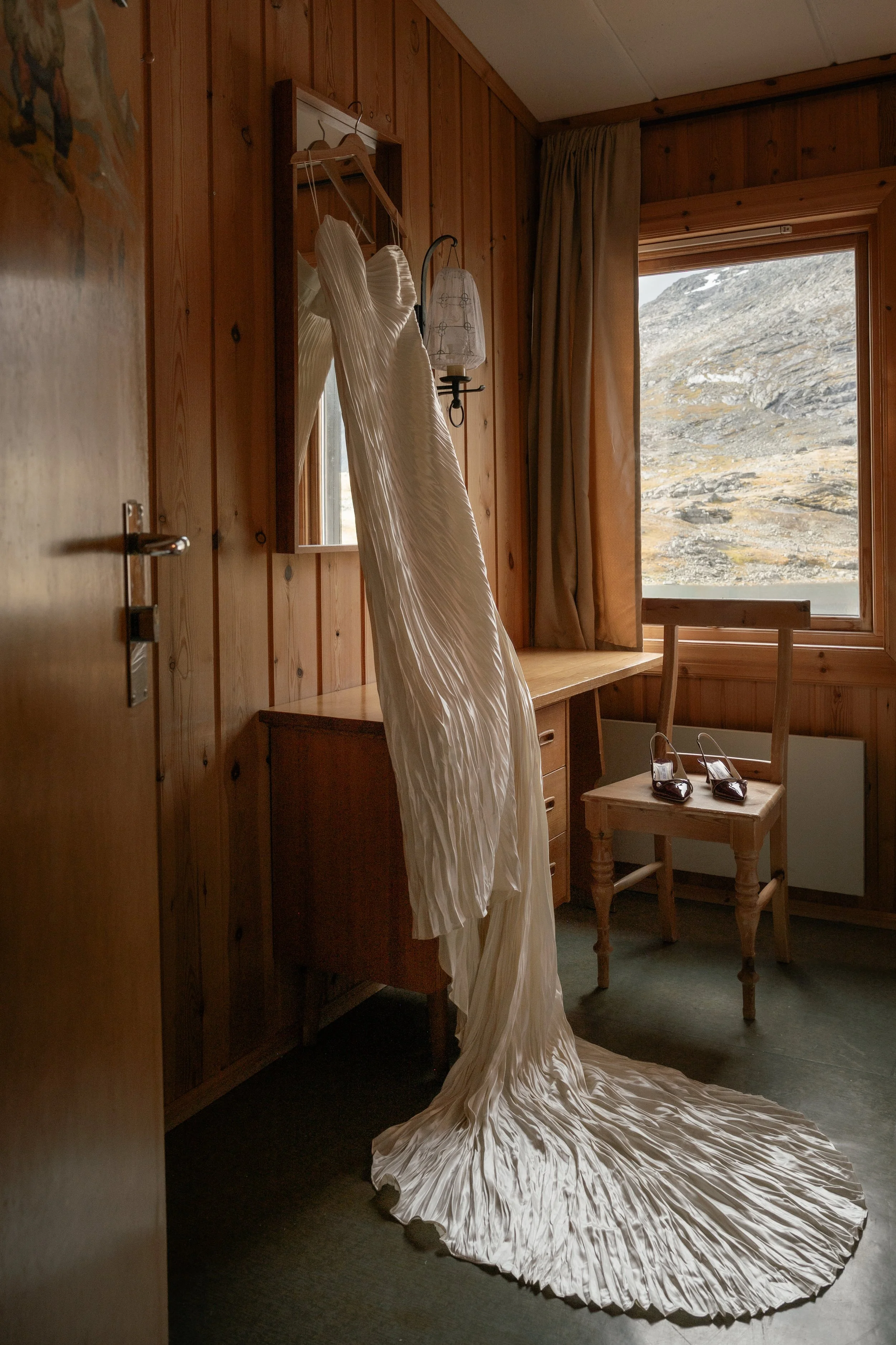 A wedding dress hanging on a mirror in a wooden-paneled room with a view of mountains outside.