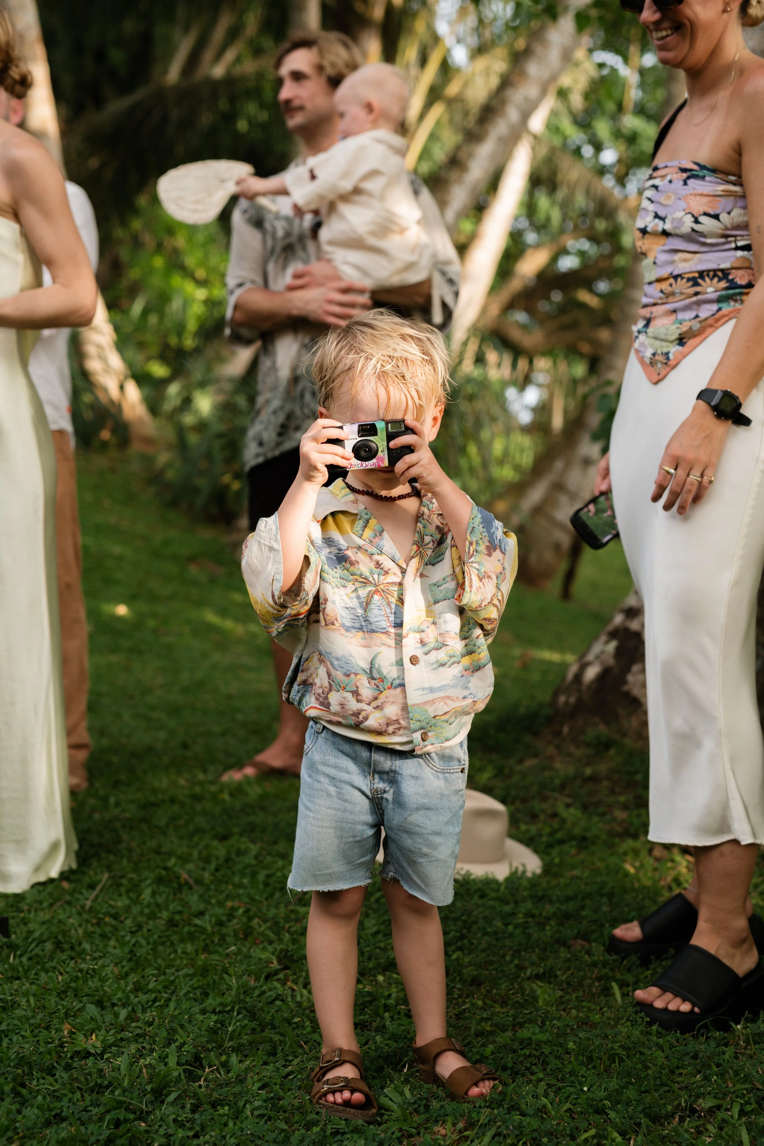 A young boy with blond hair and a tropical shirt is taking a picture with a vintage camera in an outdoor setting with trees and grass, surrounded by adults.