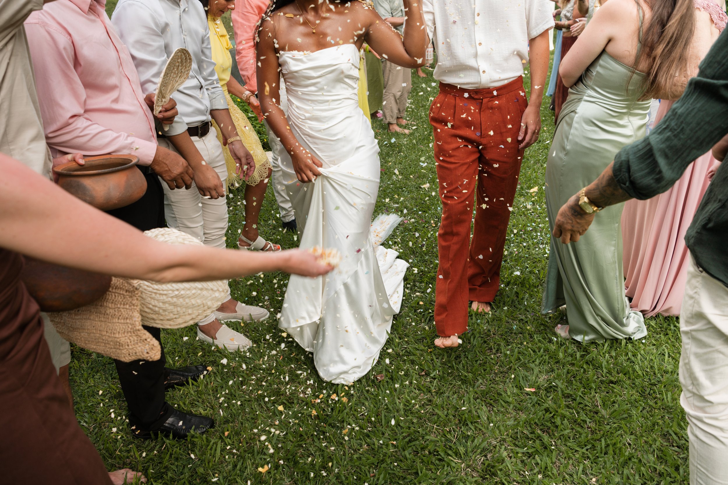 People at a wedding celebration outdoors, with a woman in a white wedding dress in the center, surrounded by guests throwing flower petals.