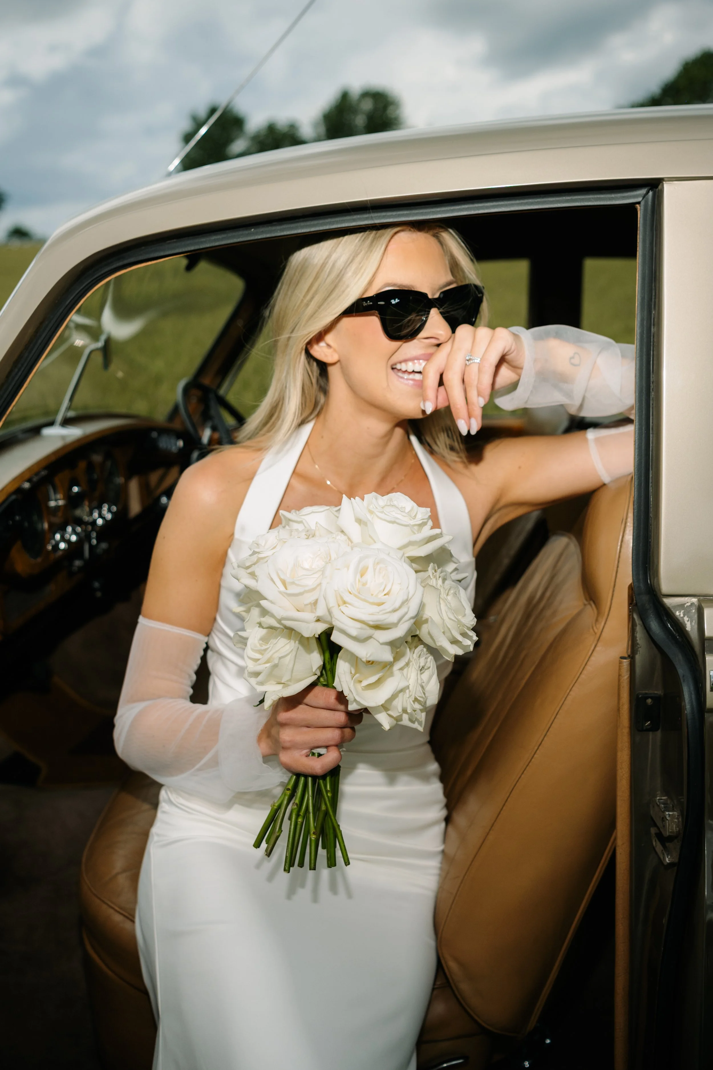 A bride sitting inside a vintage car, holding a bouquet of white roses, wearing a white wedding dress, black sunglasses, and smiling.