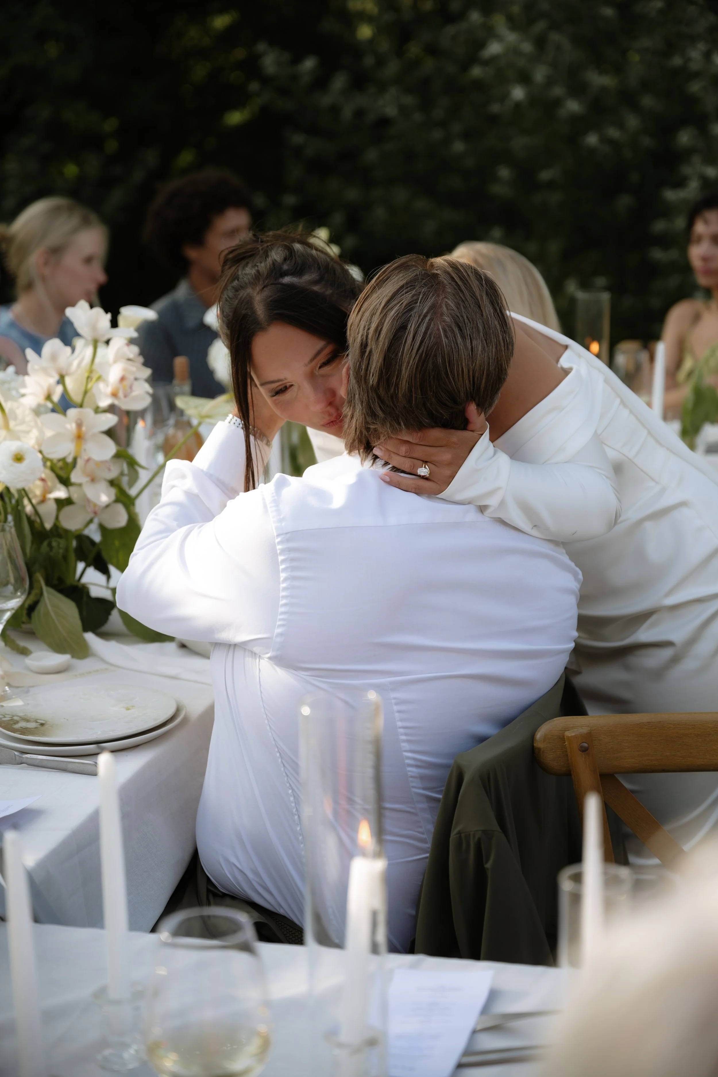 A woman in a white dress is kissing a man sitting at a table, surrounded by friends at an outdoor gathering.
