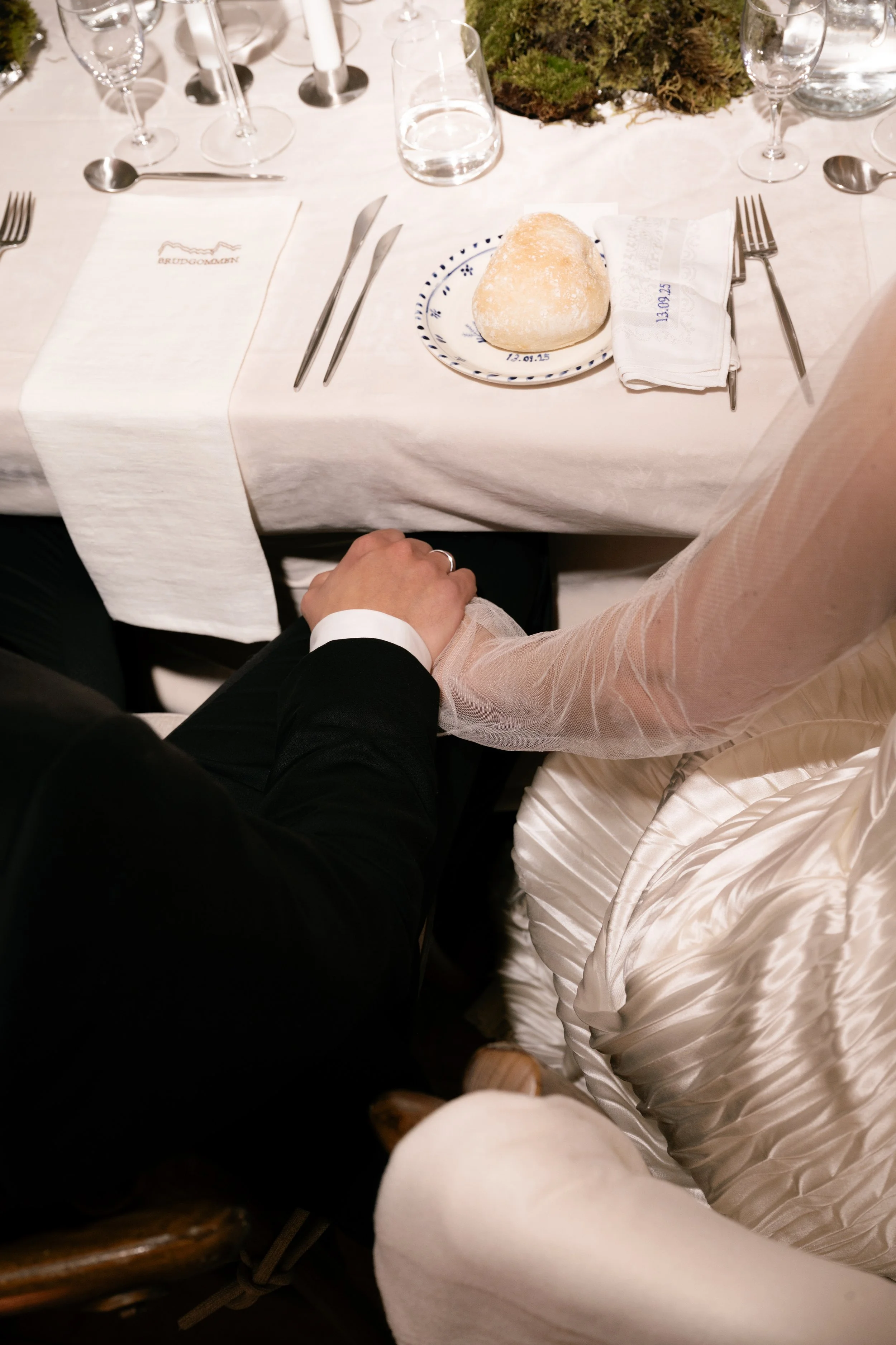 A bride and groom holding hands at a wedding reception table, which has a white tablecloth, a single bread roll on a plate, and set with silverware, glassware, and a napkin.