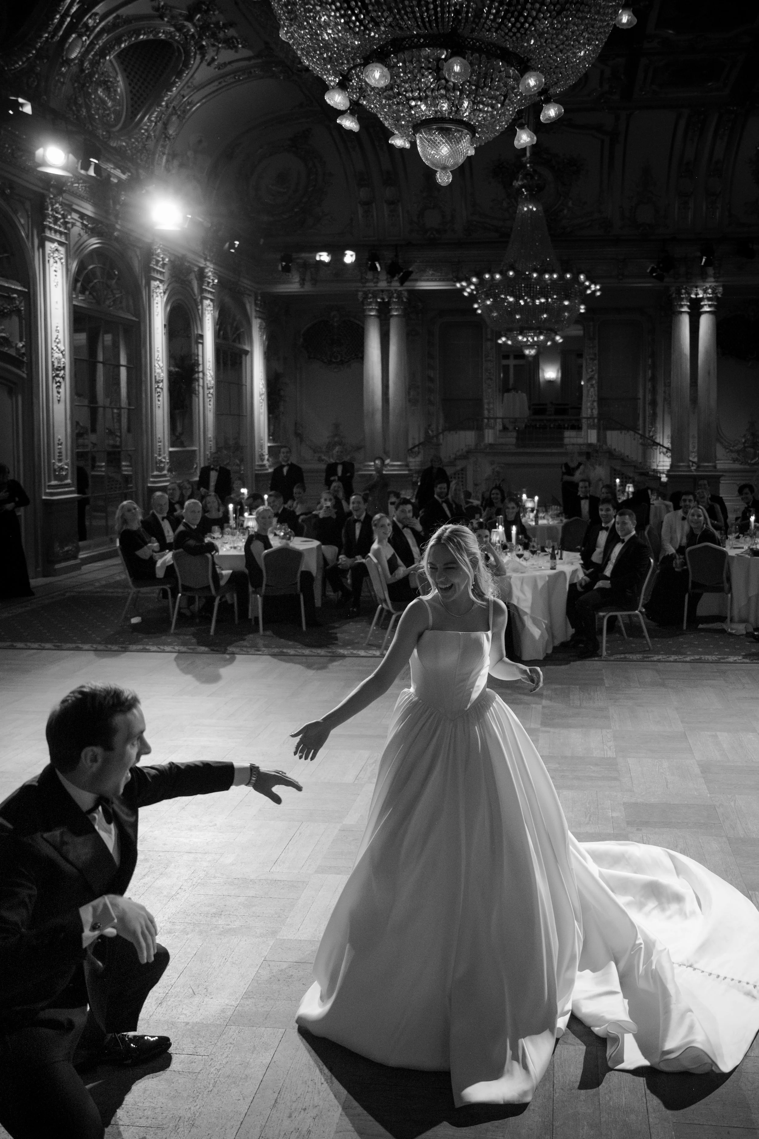 A bride dancing with a groom at a wedding reception in an ornate ballroom with chandeliers and seated guests.