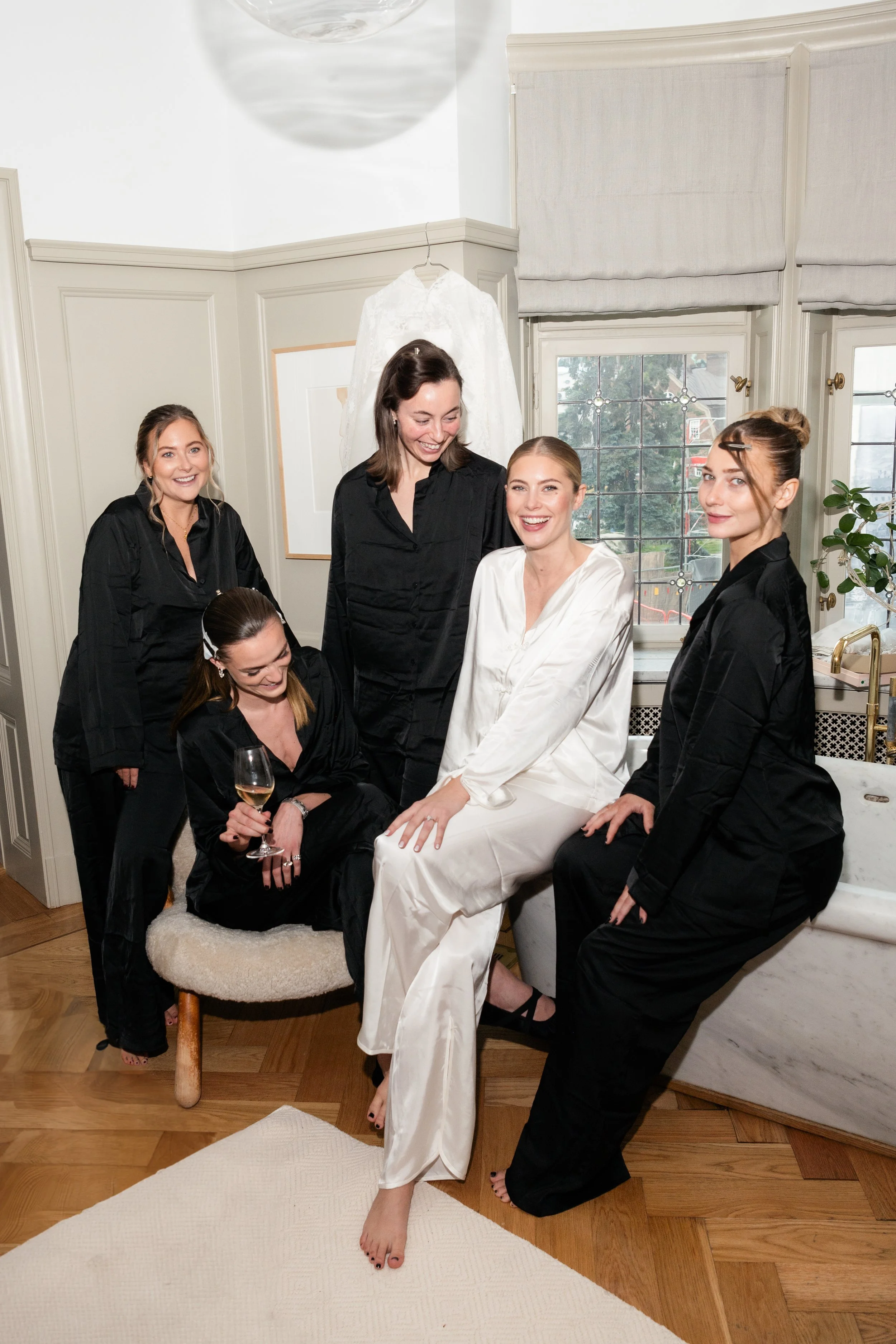 Group of women, with one in a white satin dress, laughing and smiling in a well-lit room with large windows and light-colored walls, surrounded by four women in black pajamas.
