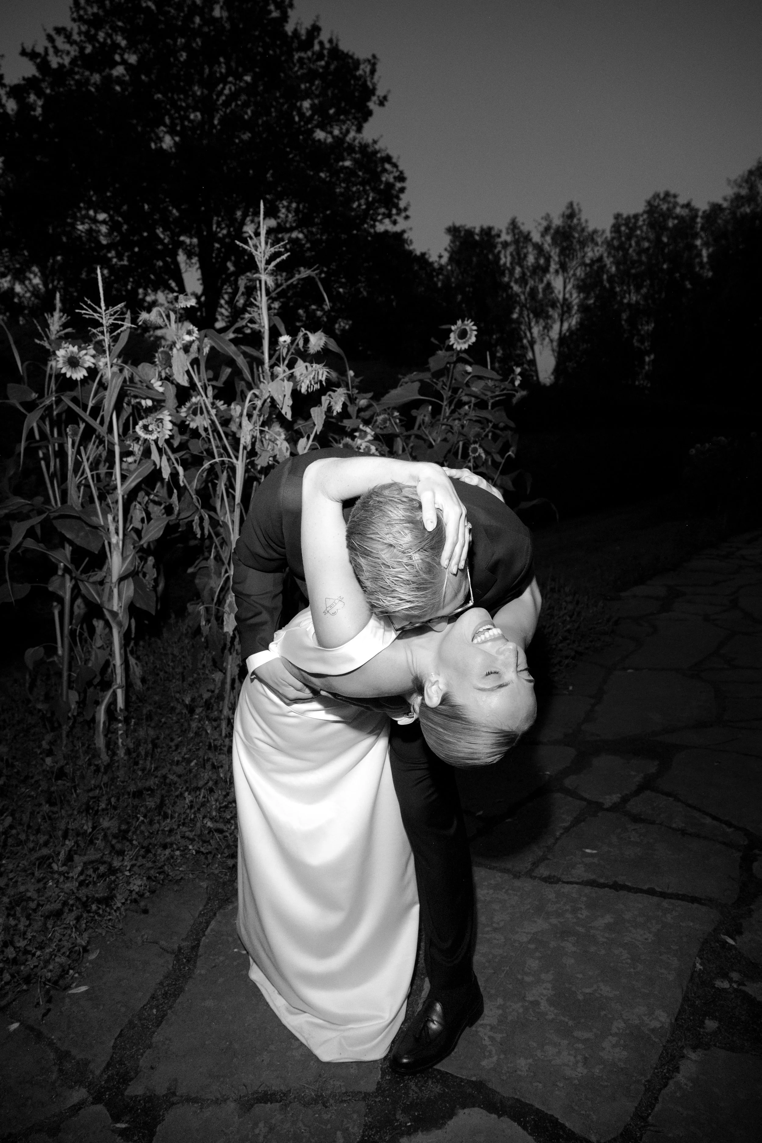 A couple dressed in wedding attire sharing a joyful kiss outdoors at night, with the groom dipping the bride and a row of sunflowers in the background.
