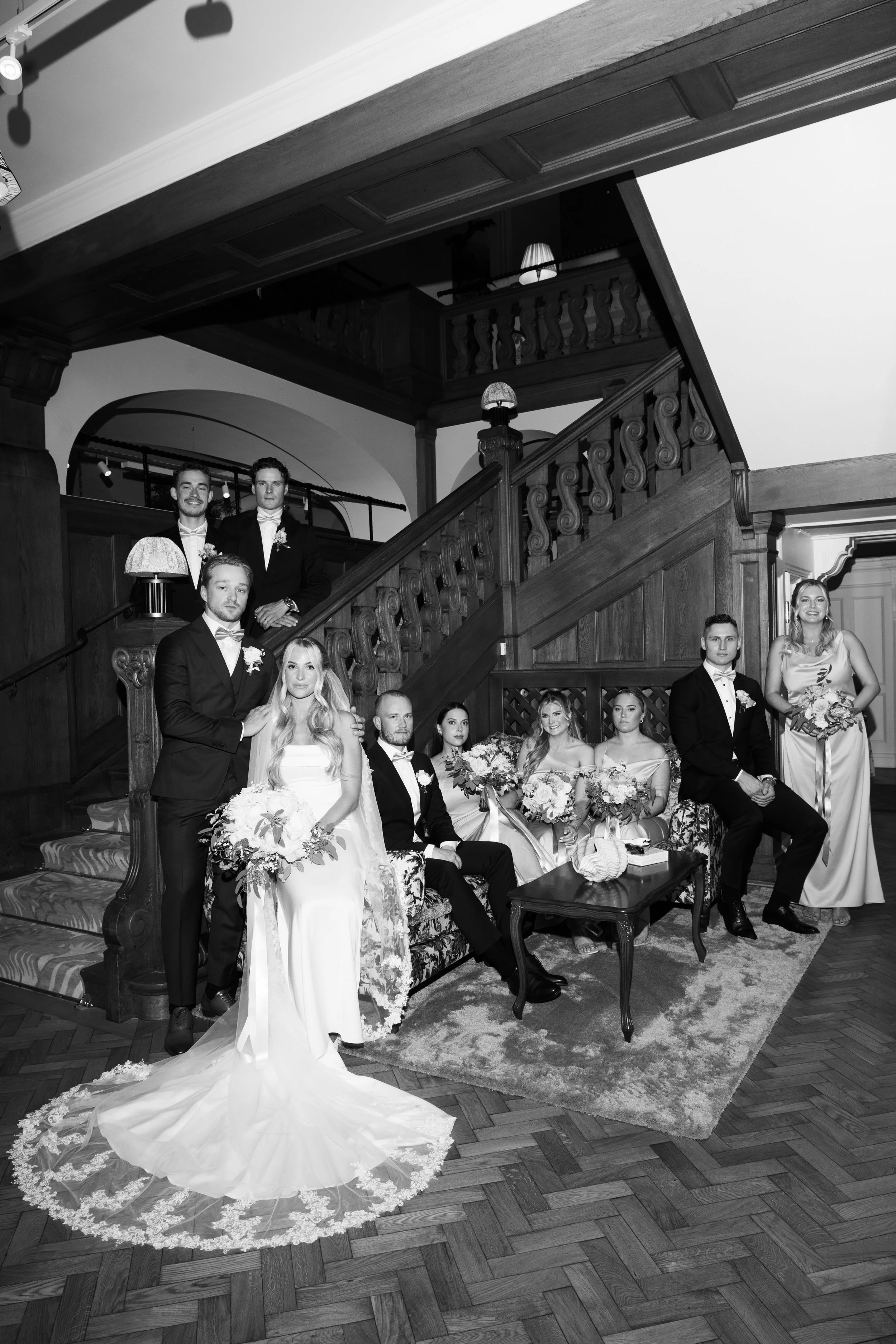 A black-and-white photo of a wedding party with the bride, groom, bridesmaids, and groomsmen gathered in a fancy indoor setting with wooden staircase and elegant furniture.