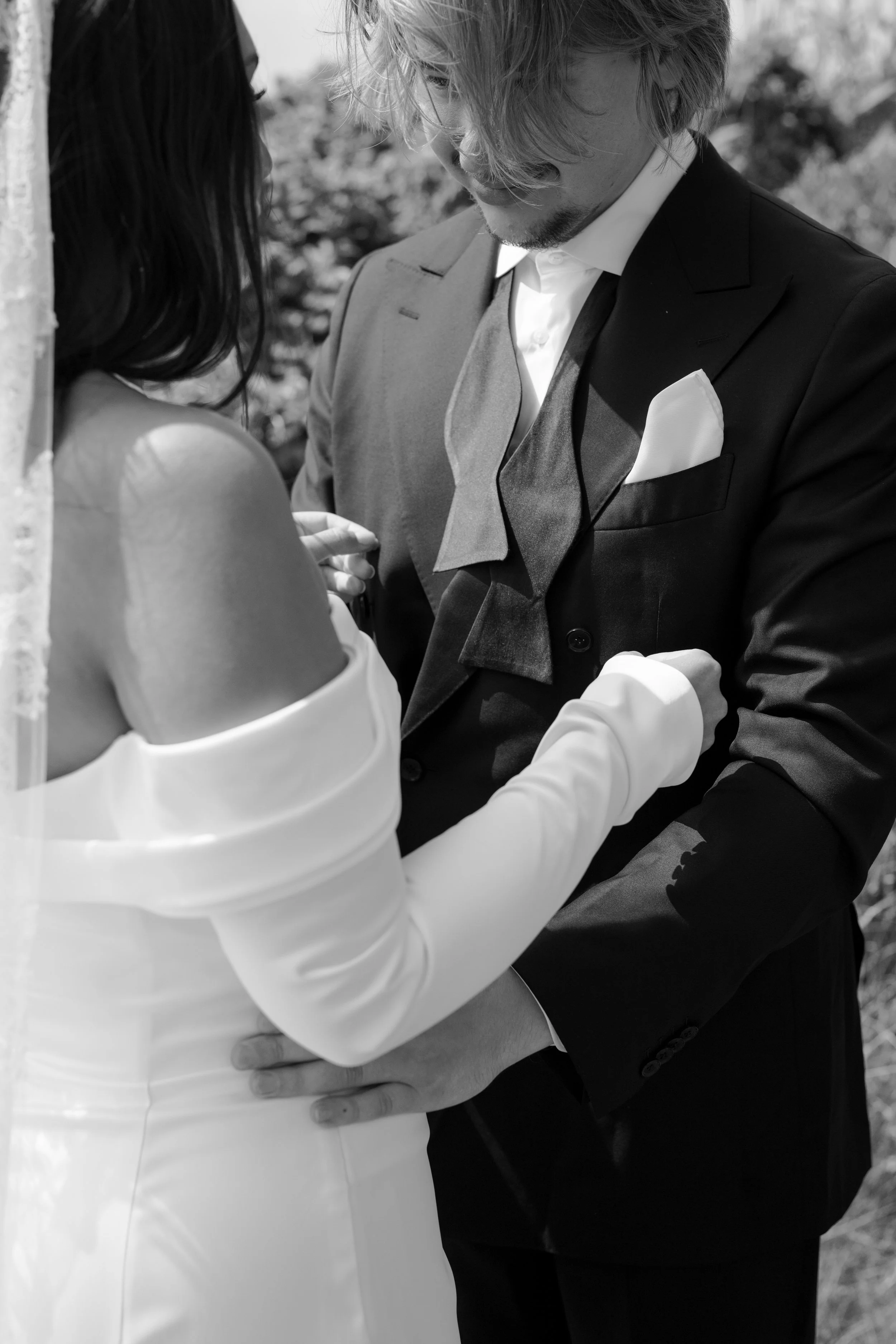 A couple dressed in formal wedding attire embrace outdoors, with the man tenderly holding the woman's waist.