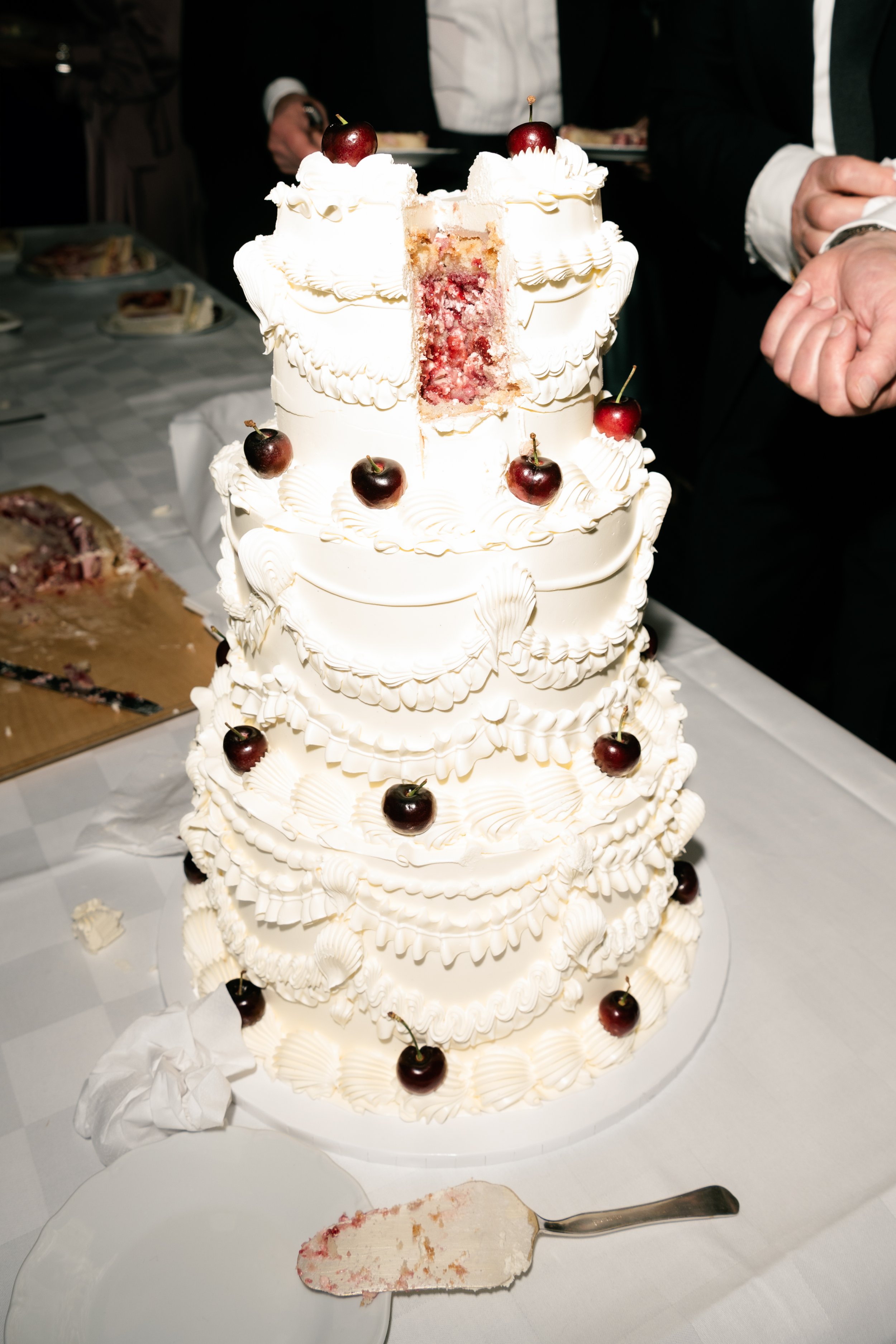 A large white wedding cake with multiple tiers decorated with cherries and white frosting, partially sliced to reveal pink filling inside.
