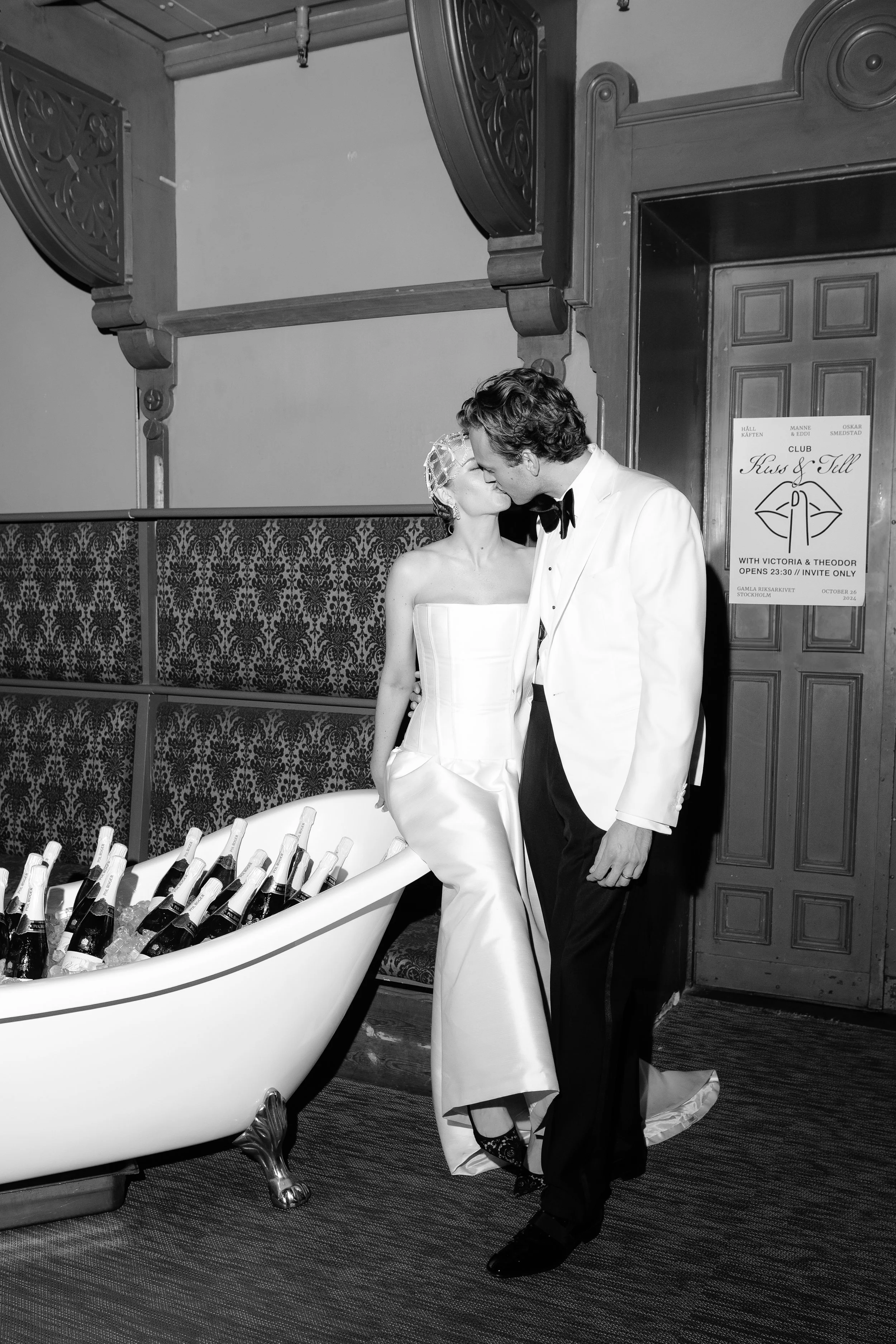 A bride and groom share a kiss at their wedding reception, standing next to a bathtub filled with bottles of champagne.