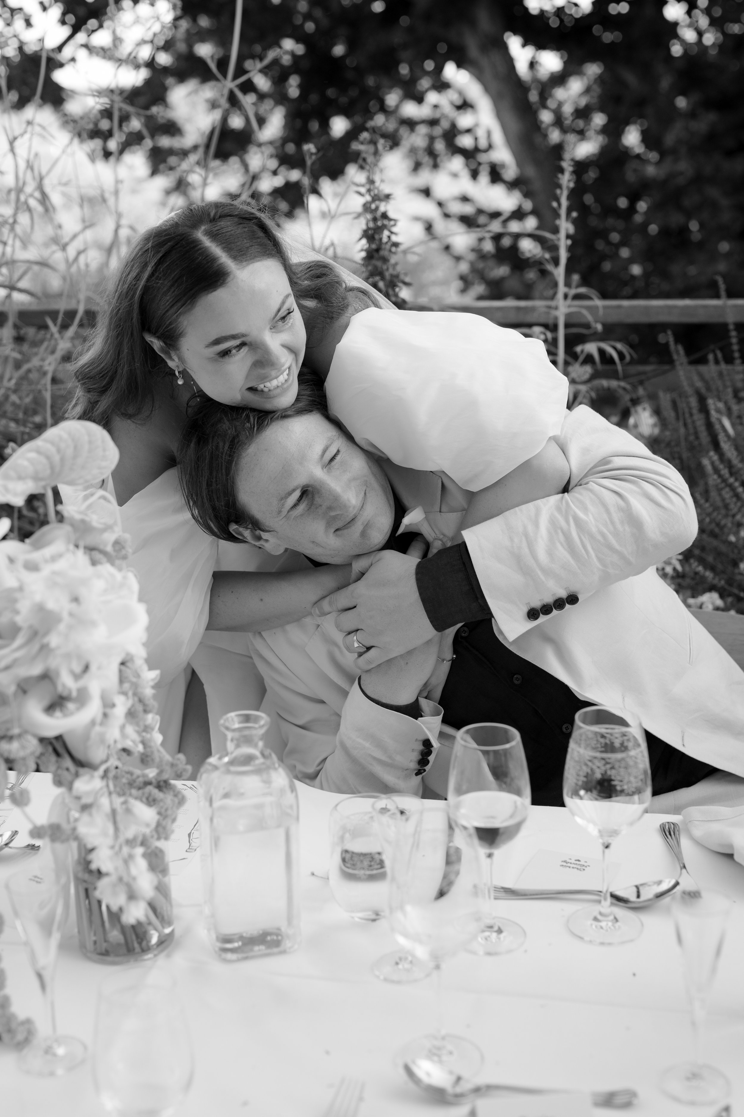 A black and white photo of a joyful moment at a celebration, showing a woman and two men hugging and smiling amid a decorated table with glasses and a floral arrangement, outdoors with trees in the background.