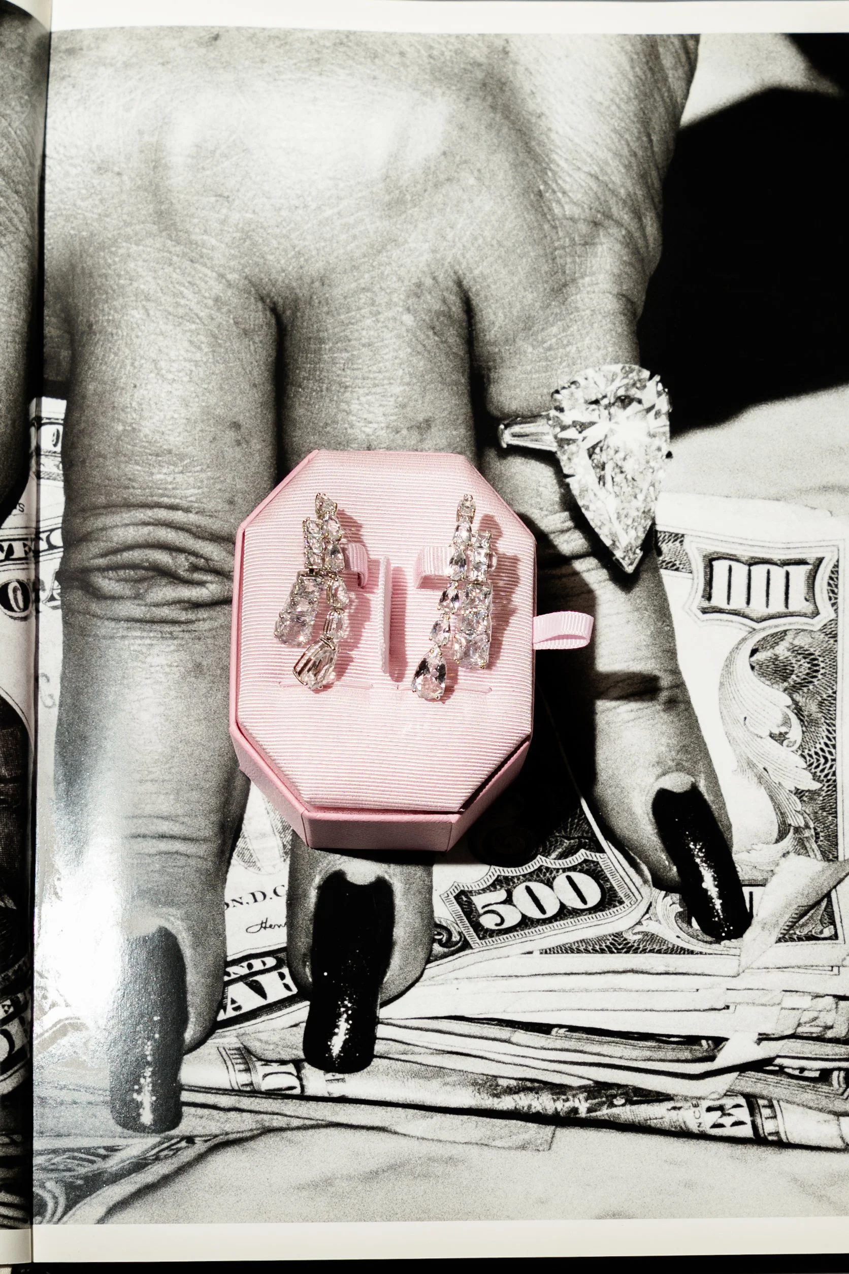 A black and white photograph of a hand with black painted nails and a large diamond engagement ring, with jewelry displayed on top of a pink jewelry box resting on the hand.