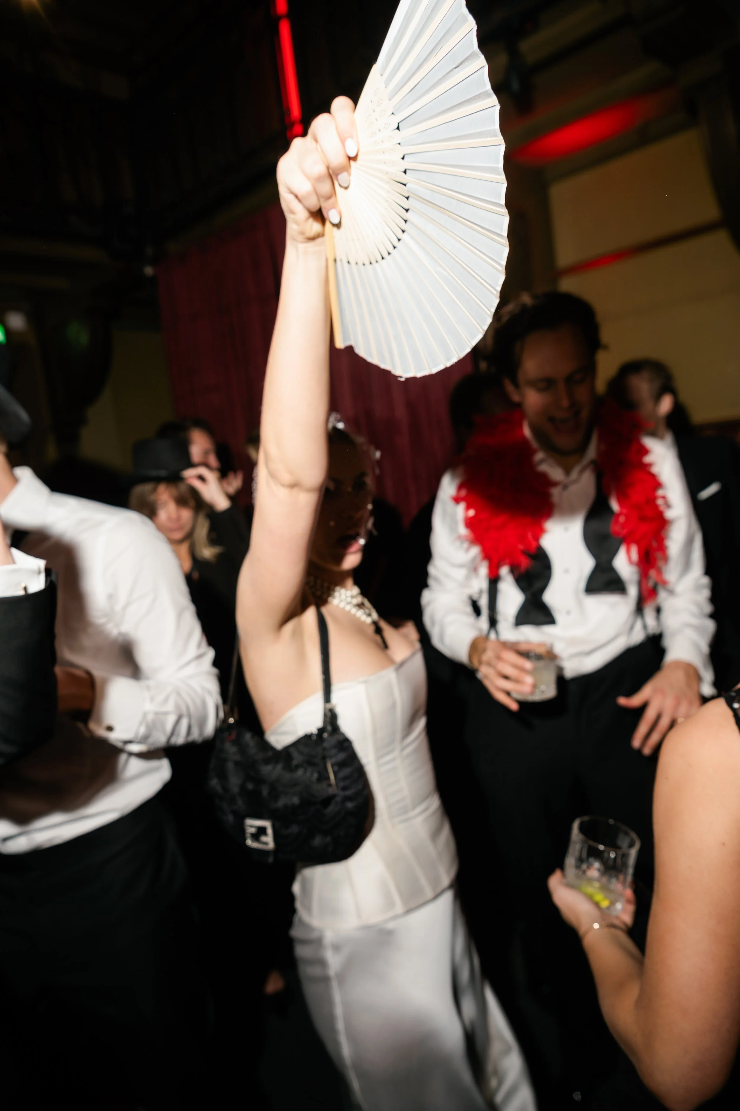 A woman holding a handheld fan above her head at a party or nightclub, with other people dancing and socializing around her.