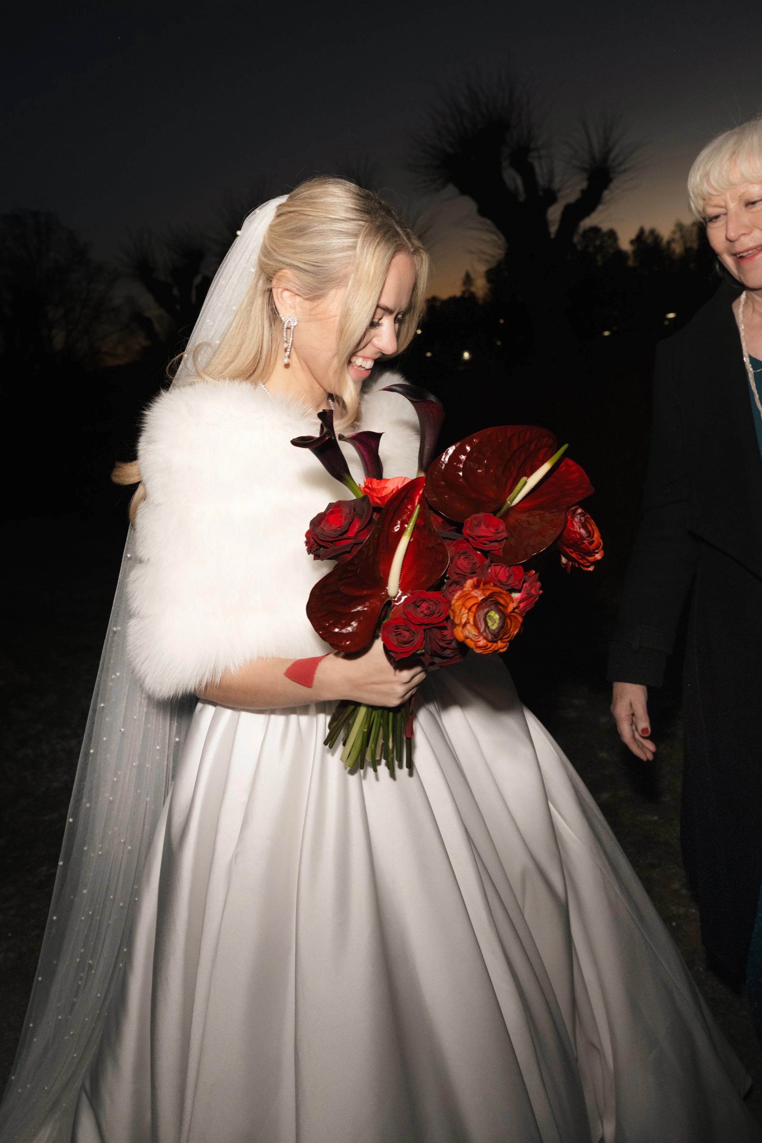 A bride in a white wedding gown holding a bouquet of red and dark purple flowers, smiling, during an outdoor evening wedding.