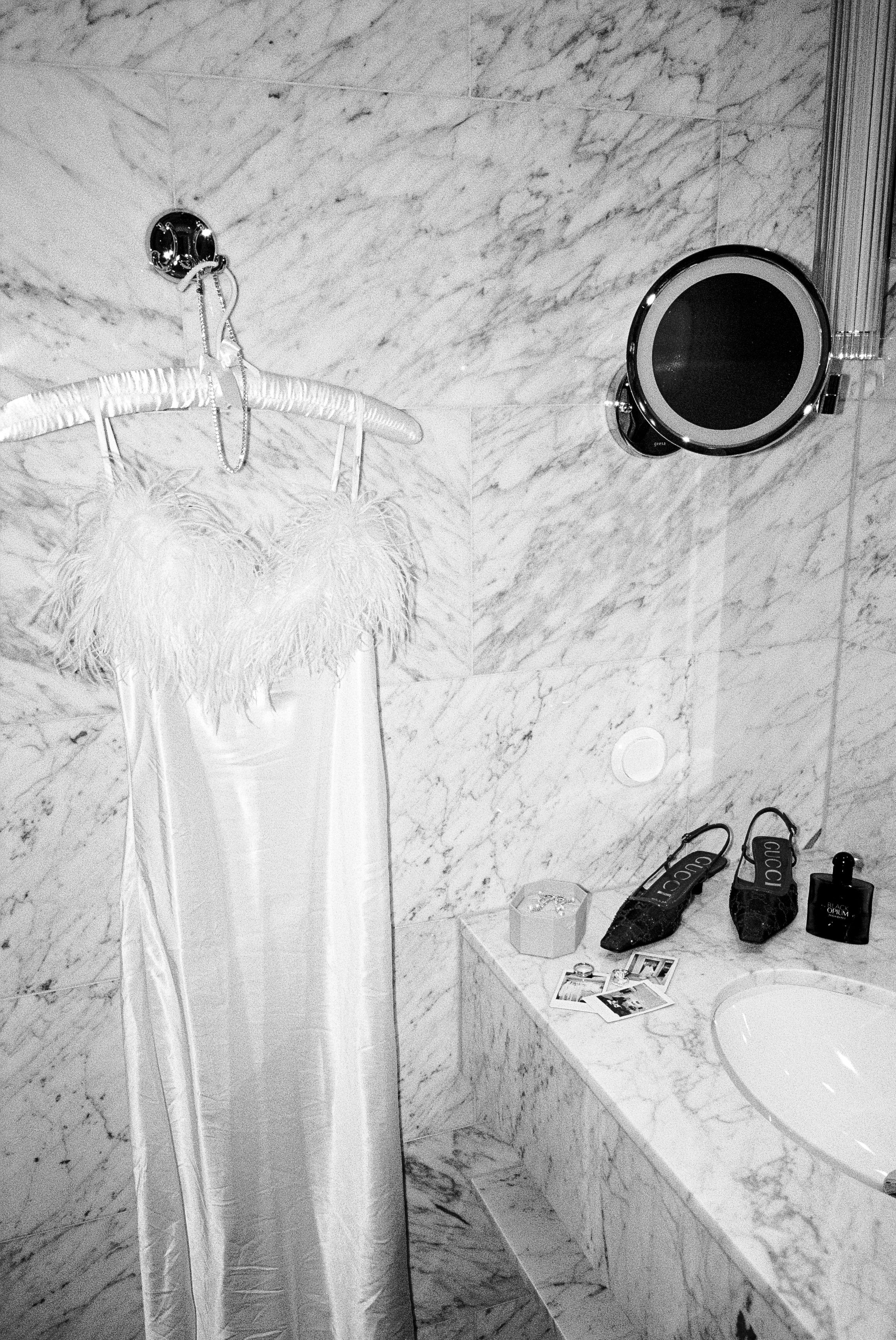 A marble bathroom wall with a hanging feather-trimmed satin garment, a mirror, photos, and personal items on the counter.