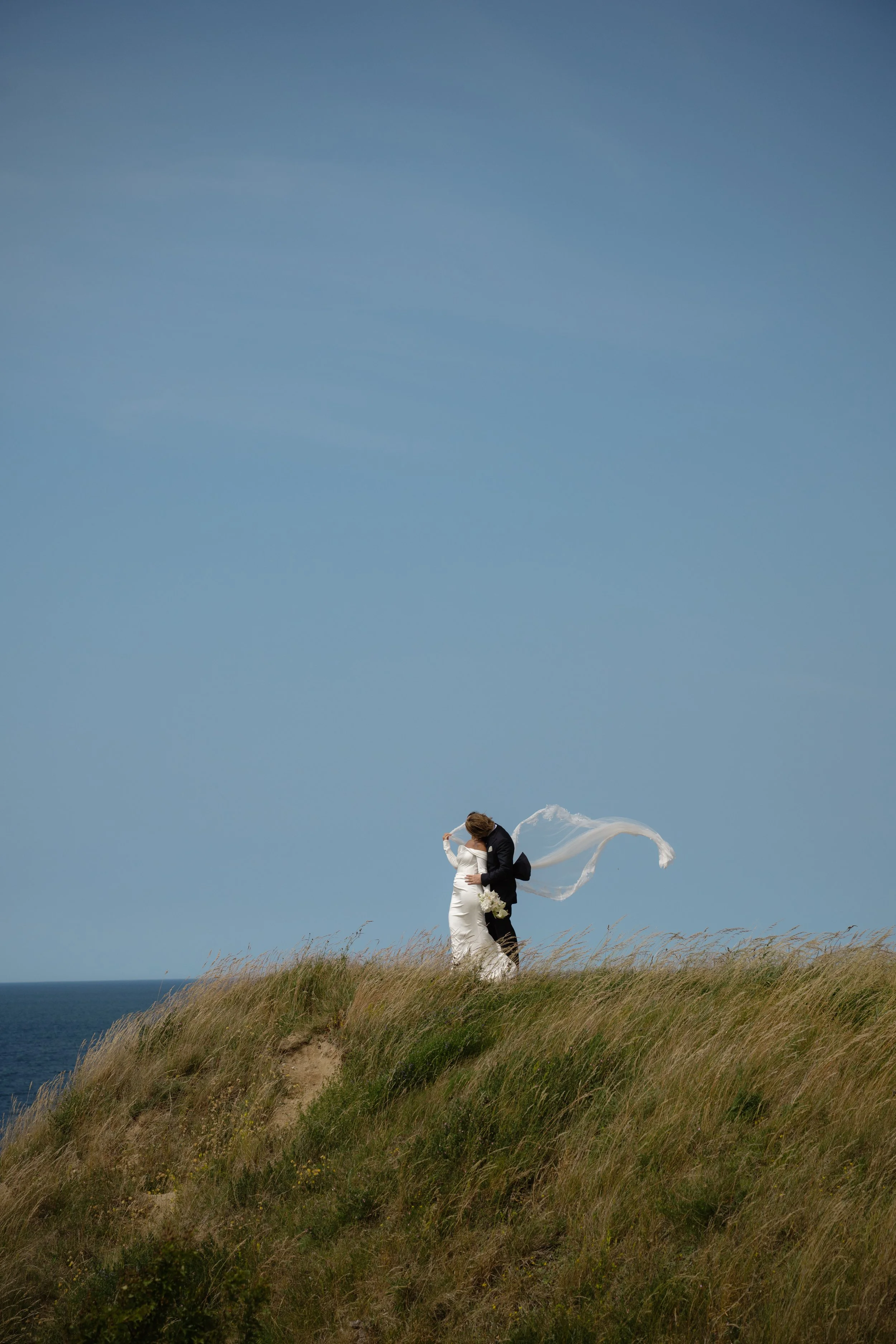 A bride and groom embracing on a grassy hilltop overlooking the ocean on a clear day.