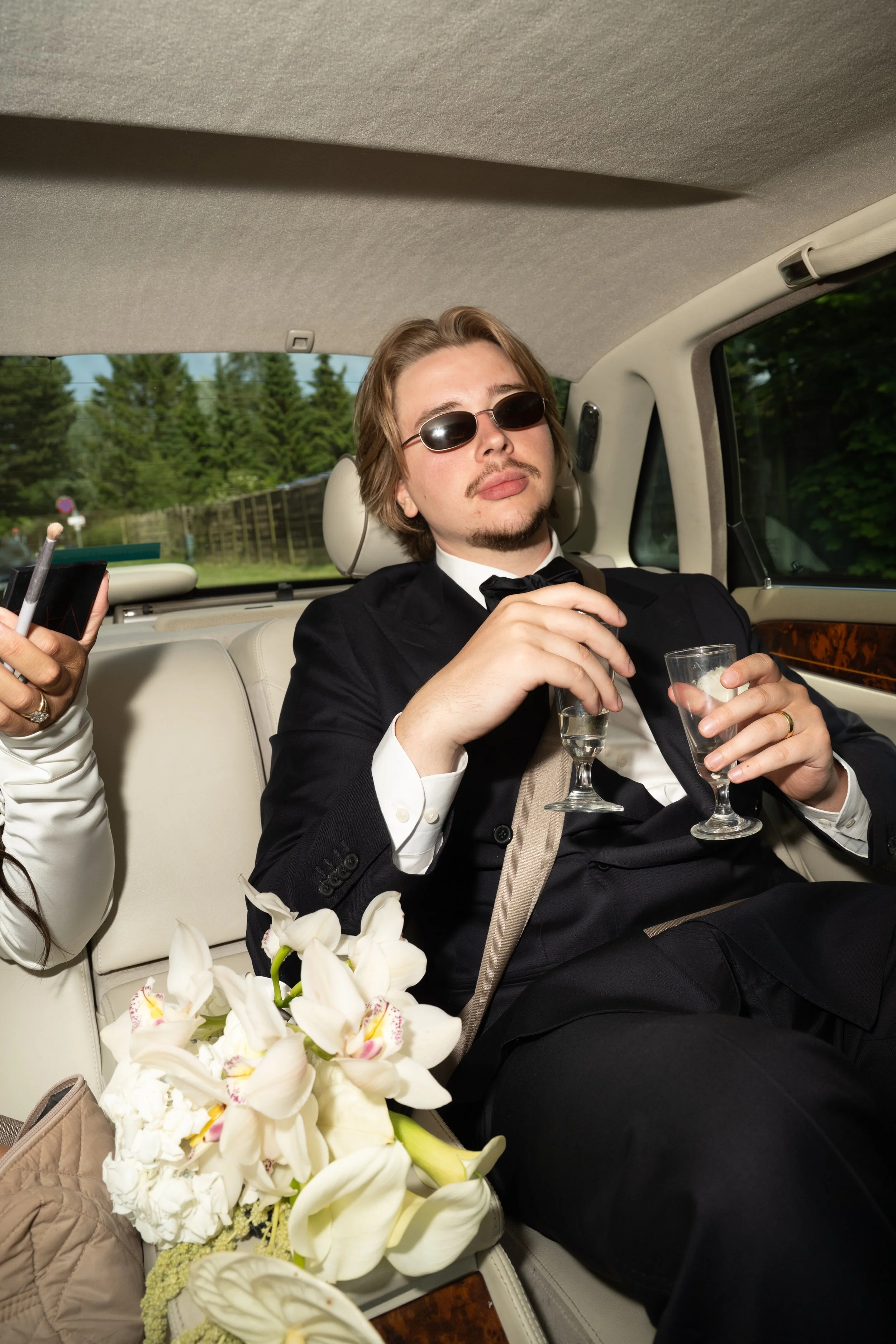 A man in a tuxedo and sunglasses sitting in the back of a car, holding two glasses of clear drinks, with white flowers on the seat beside him.