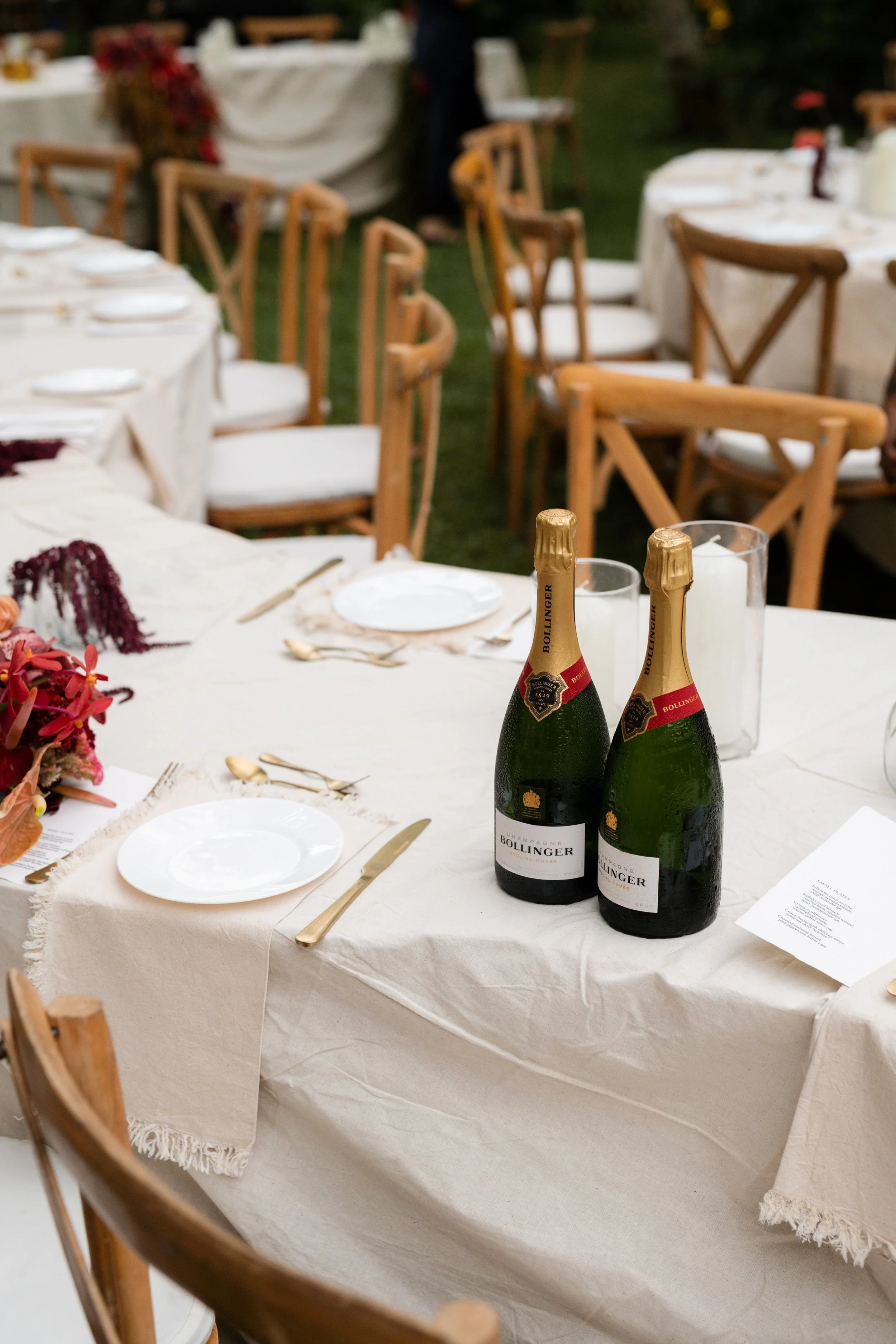 Wedding or celebration table set outdoors with champagne bottles, white tablecloths, gold utensils, and floral decorations.
