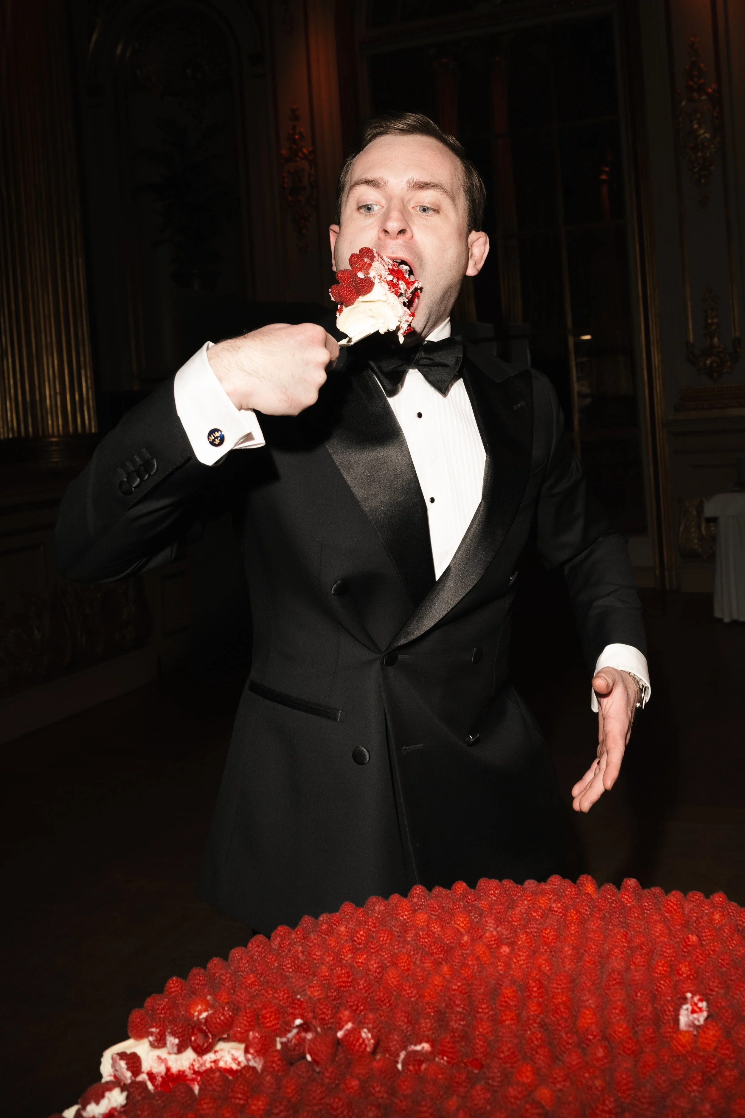 A man in a tuxedo eating a slice of cake decorated with red berries at an elegant event.