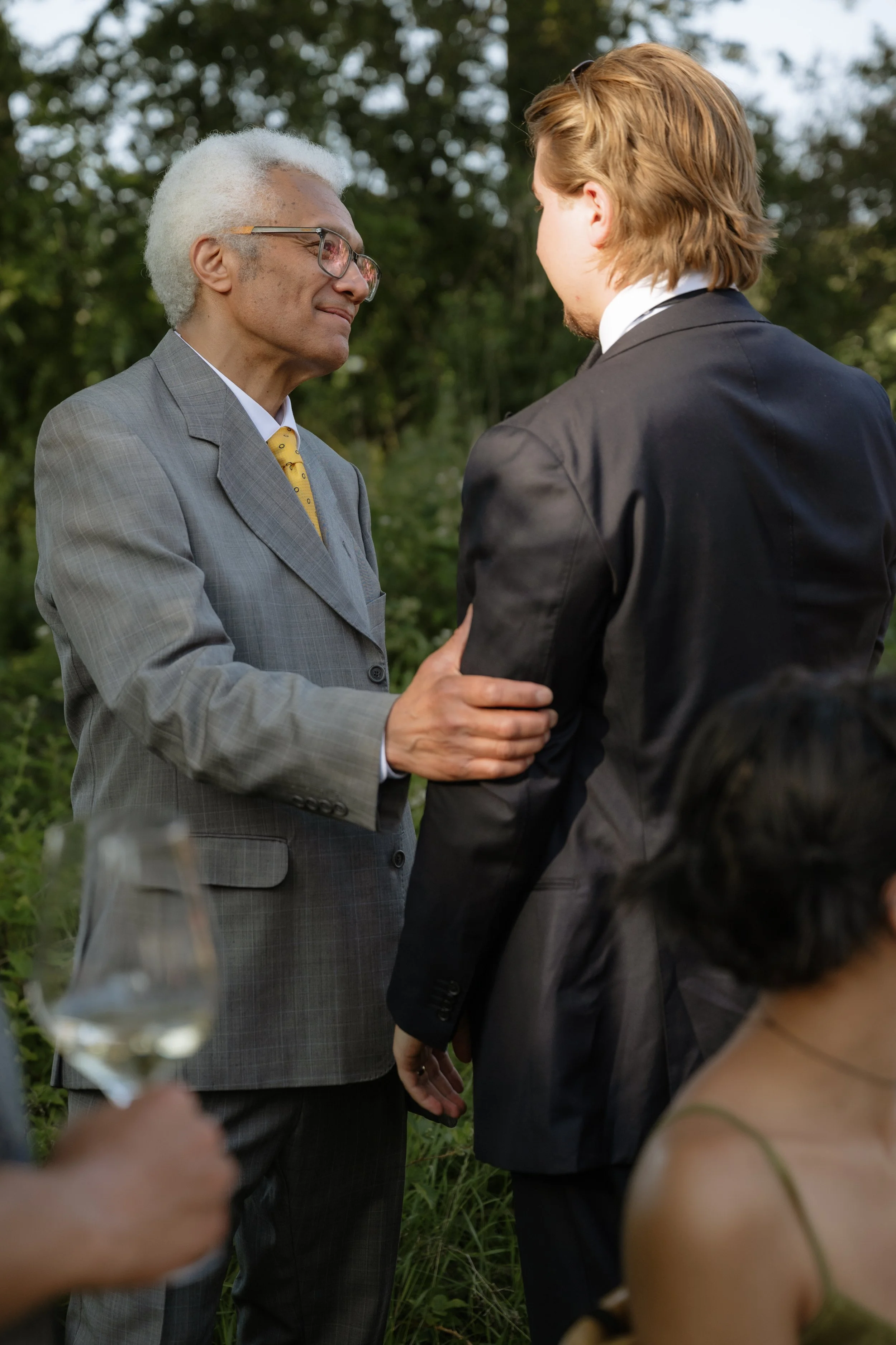 An elderly man in a gray suit and glasses is holding the arm of a younger man in a black suit during an outdoor gathering.