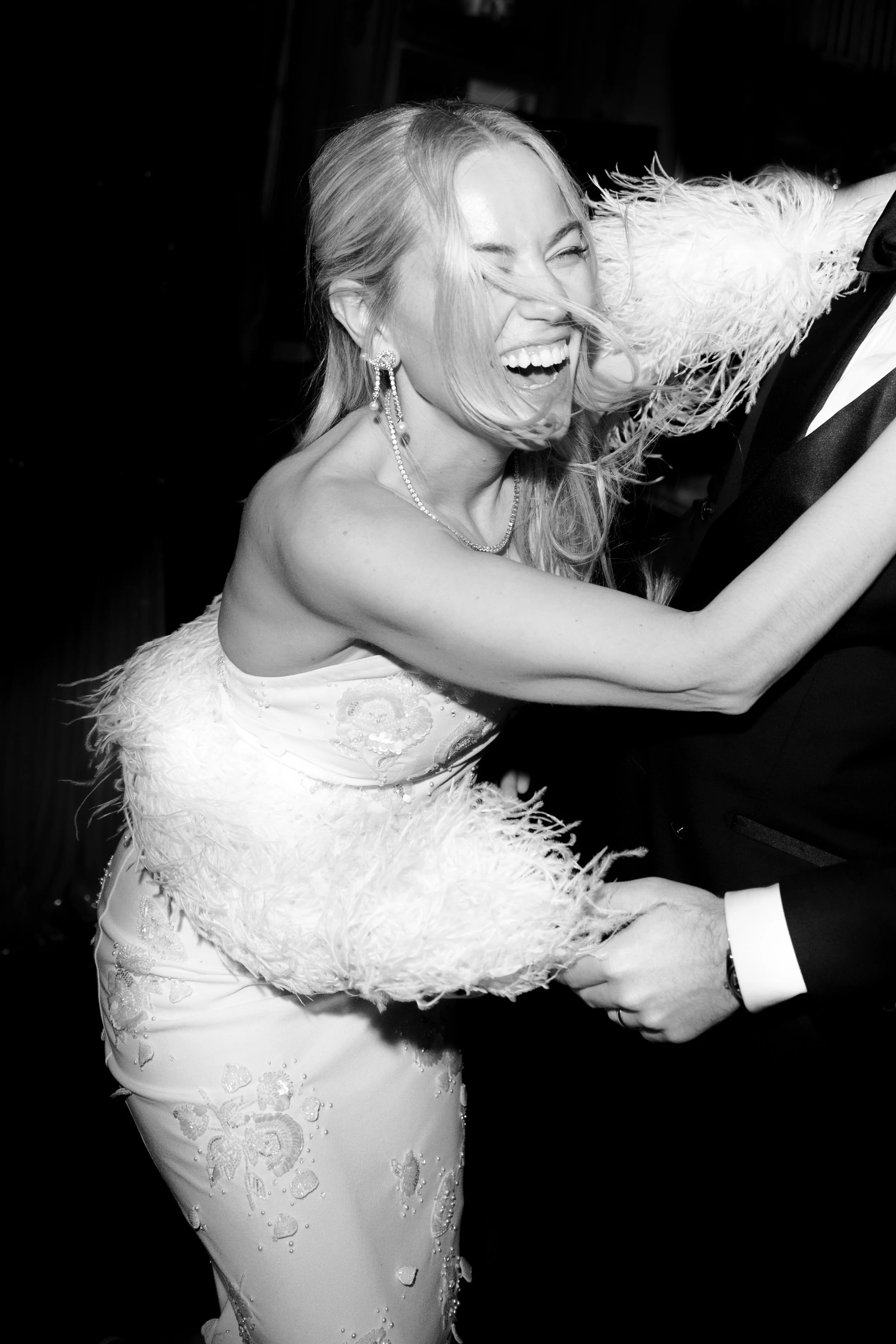 A woman in a white dress with feather details and jewelry, smiling and dancing with a man in a tuxedo.