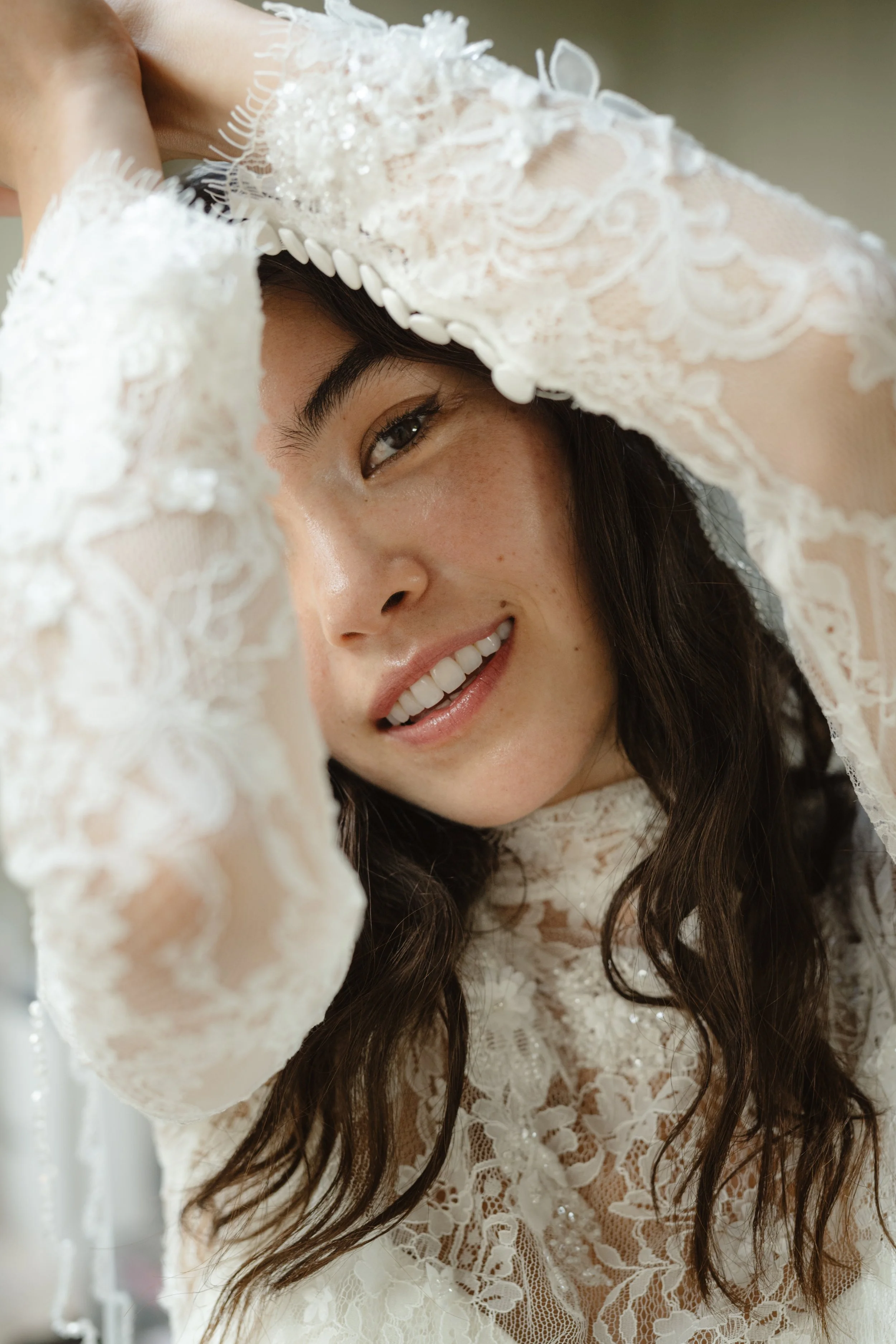 A woman smiling, wearing a lace wedding dress, holding her head with her hands, with dark wavy hair.
