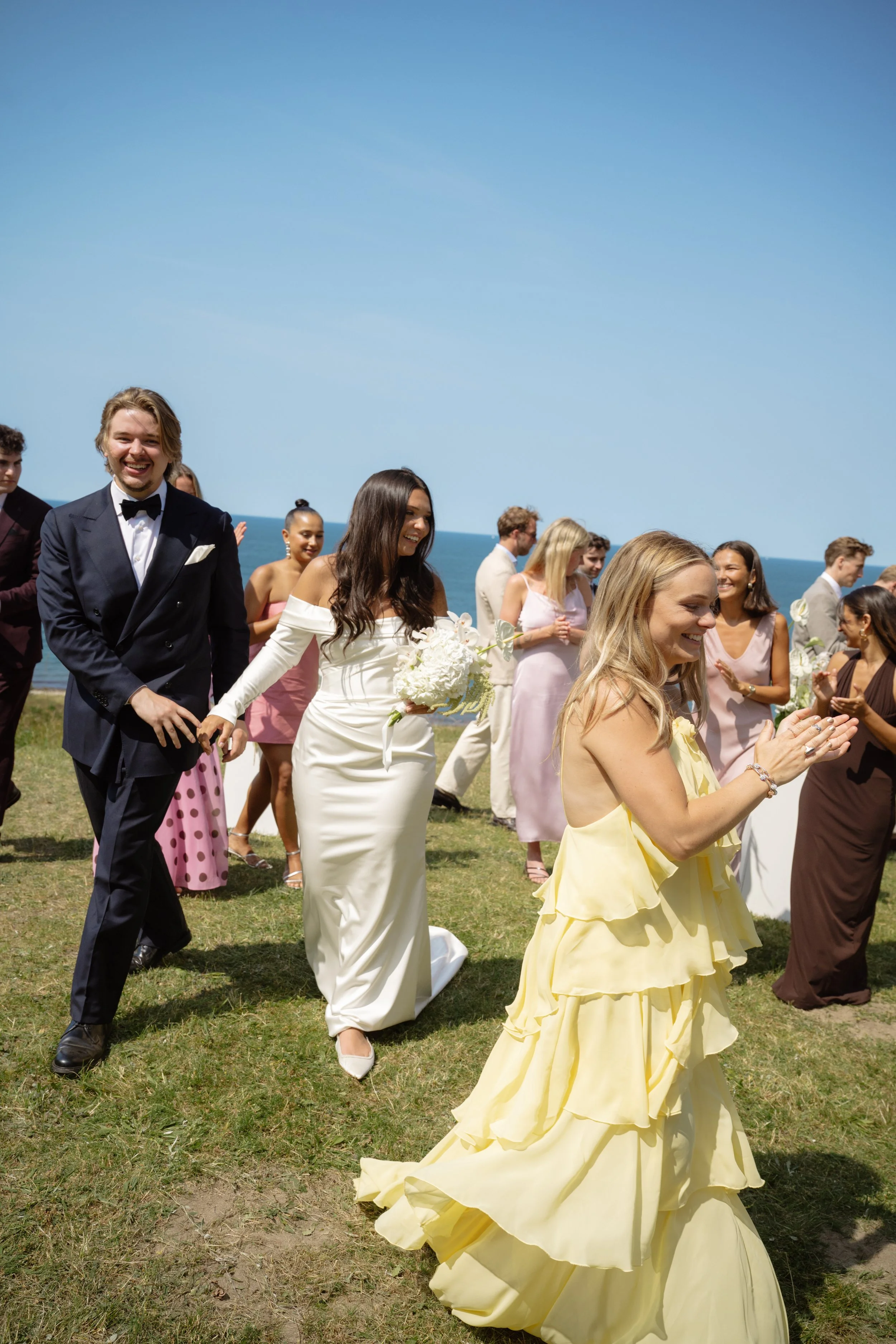 People dressed in formal attire participating in a wedding celebration outdoors near the ocean, with a clear blue sky.