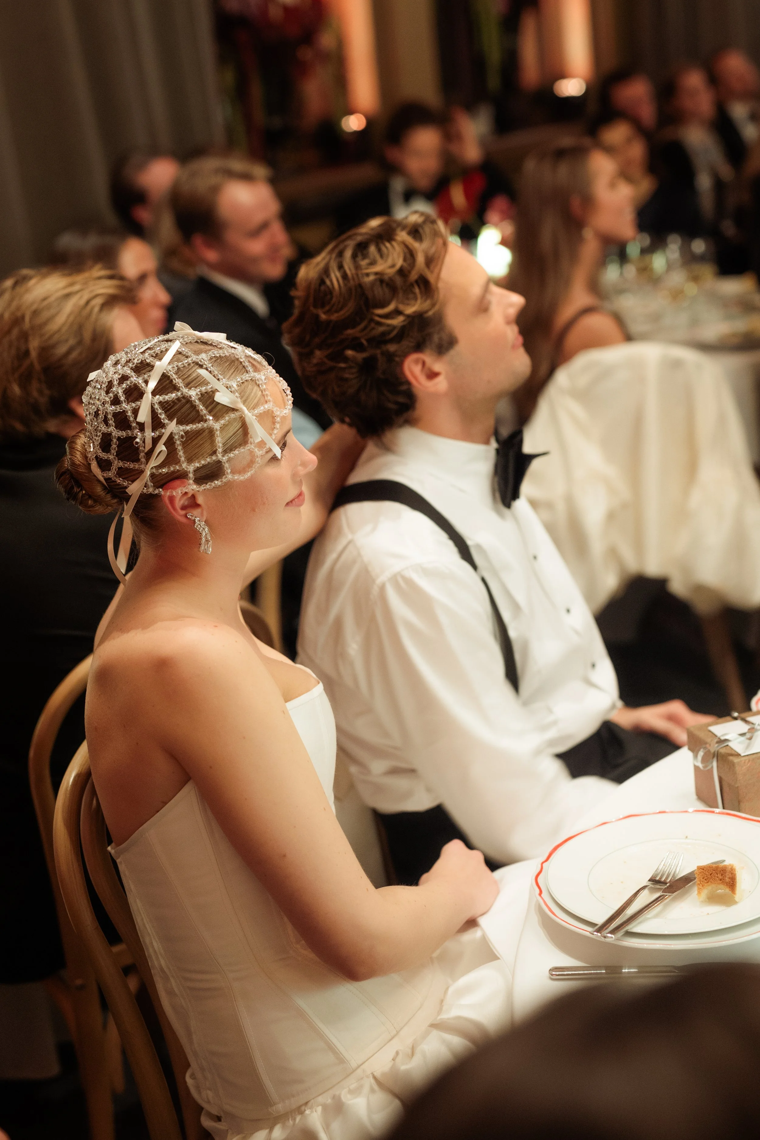 A bride and groom sit at a dinner table during a wedding reception, surrounded by guests.