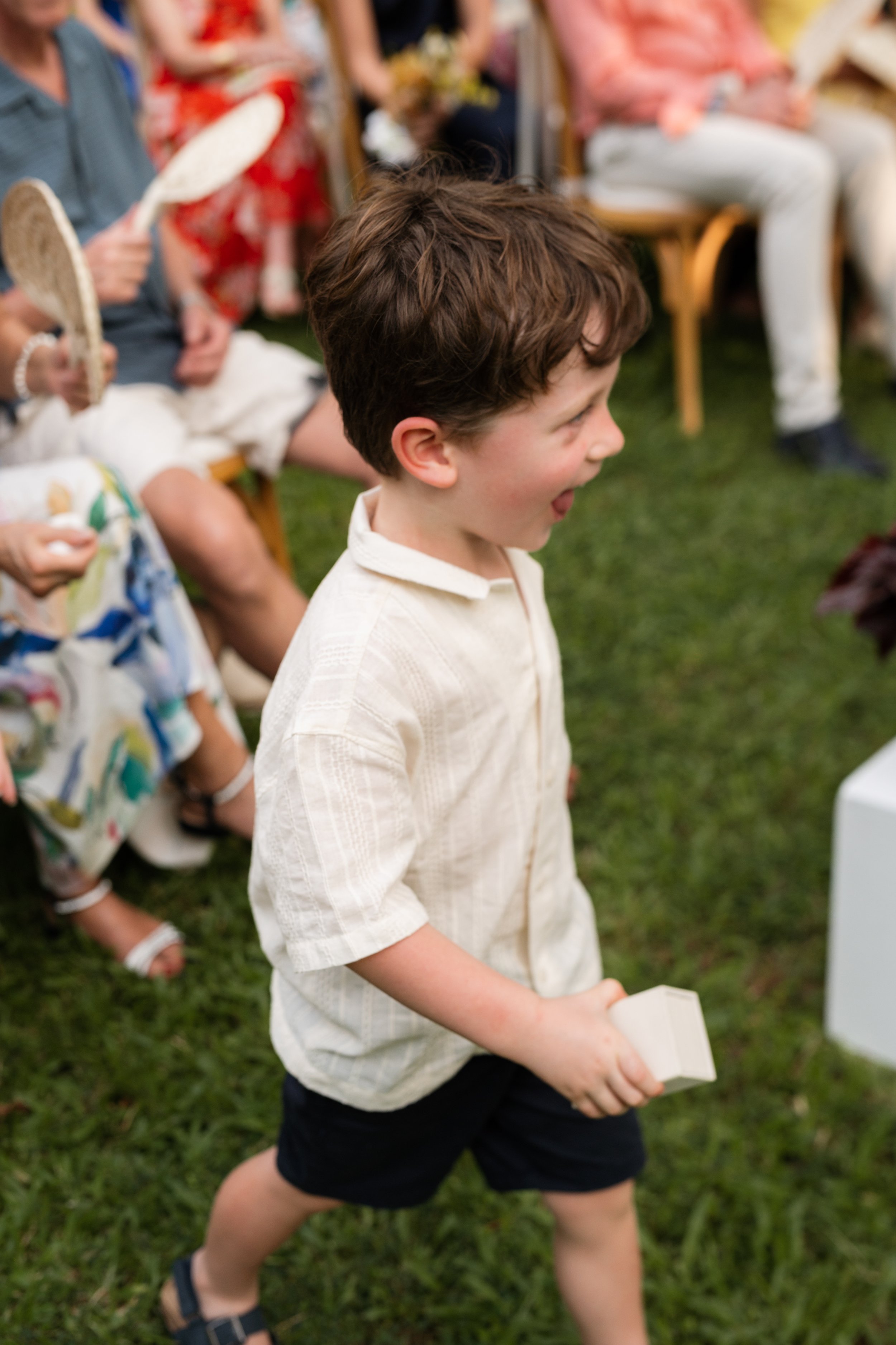 A young boy with brown curly hair wearing a cream-colored shirt and black shorts, holding a small white box, smiling and walking on grass during a celebration with other seated guests in the background.