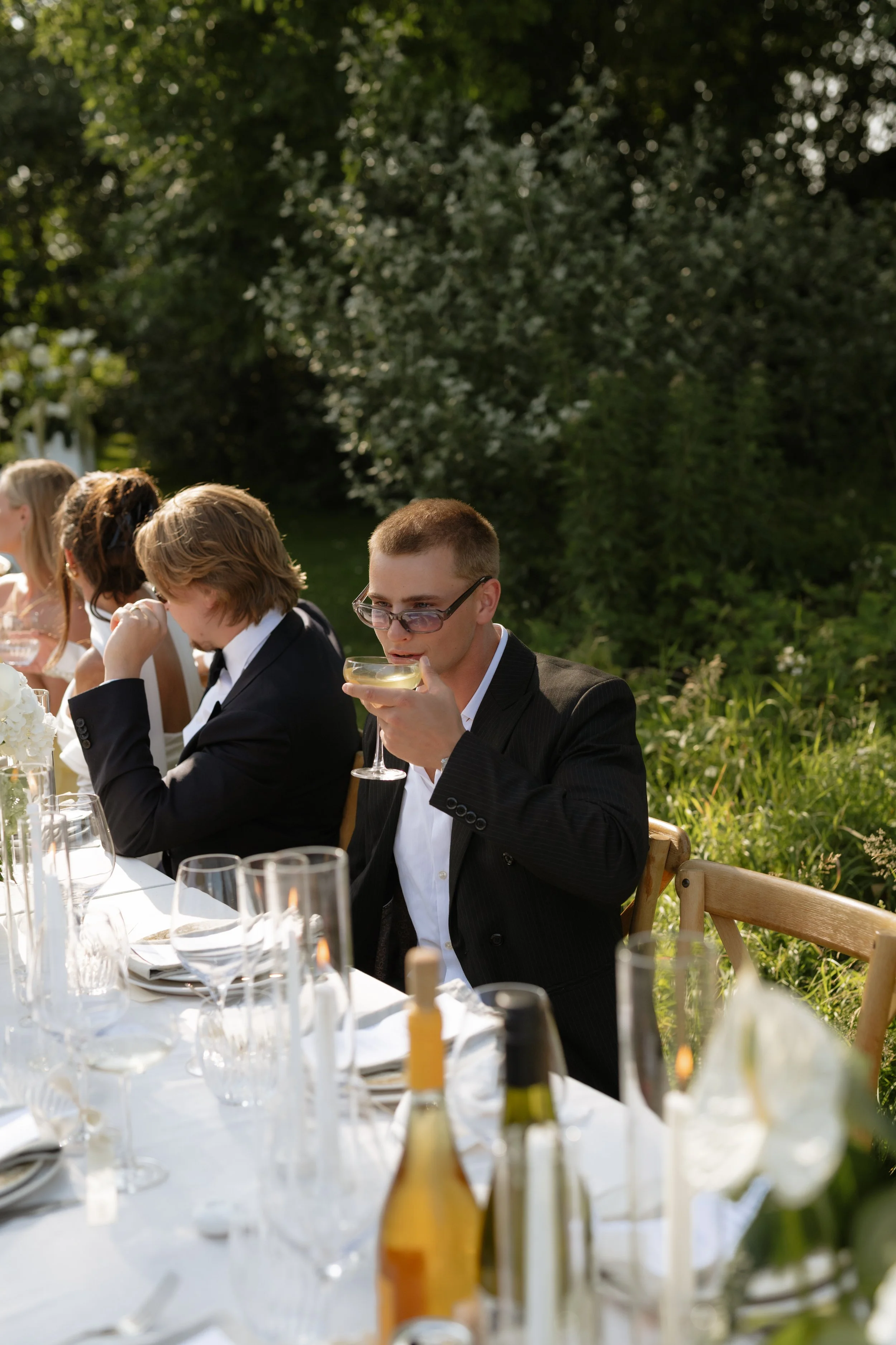 A man in a dark suit and glasses drinks wine or champagne at an outdoor dinner party with a long white table, set with glasses, bottles, and flowers, on a sunny day in a garden.