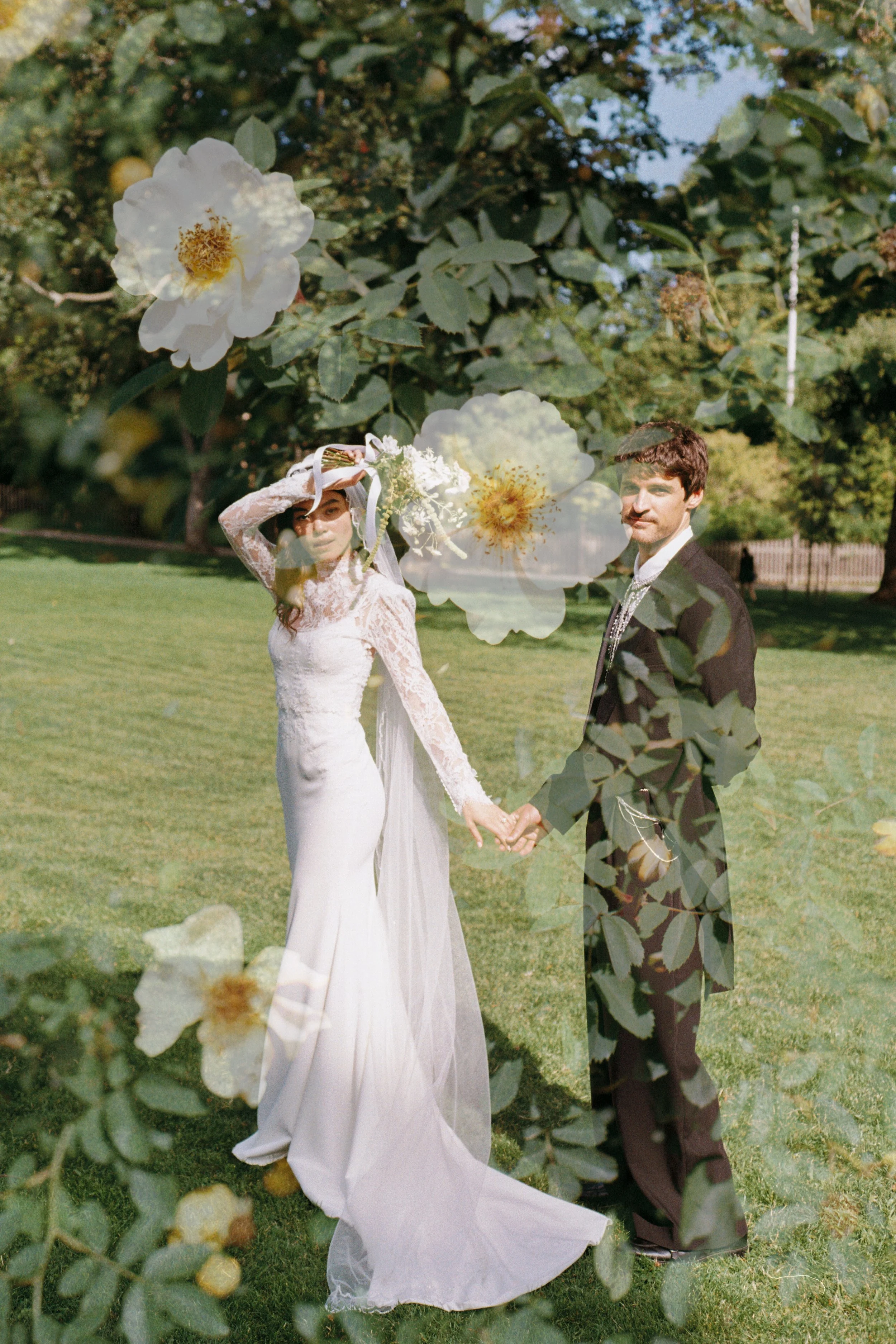 A bride and groom holding hands outdoors on a grassy area, with trees in the background. The image has a layered effect with large white flowers and leaves superimposed over the couple.
