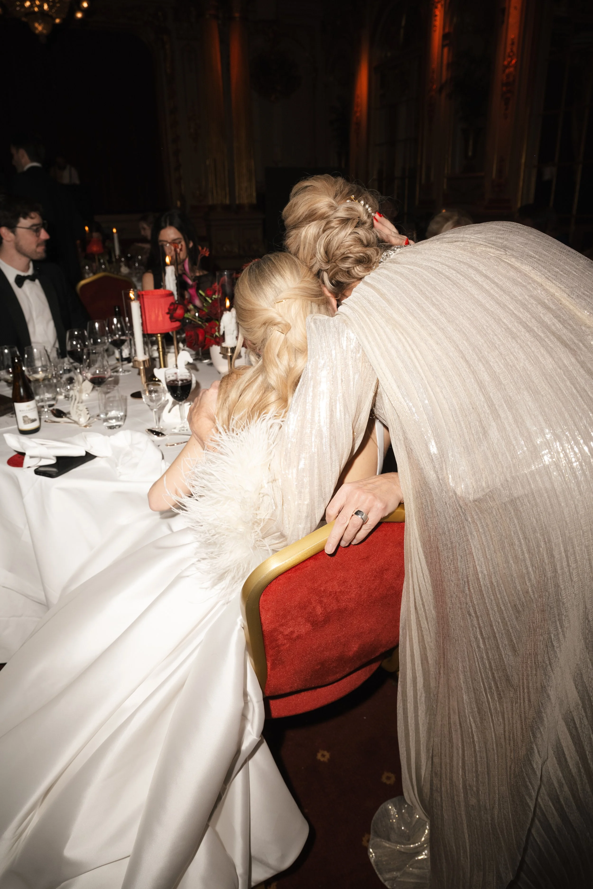An emotional moment at a formal event where a woman in a white dress is being comforted by an older woman in a light-colored gown. They are seated at a table decorated with red roses, candles, and wine glasses.