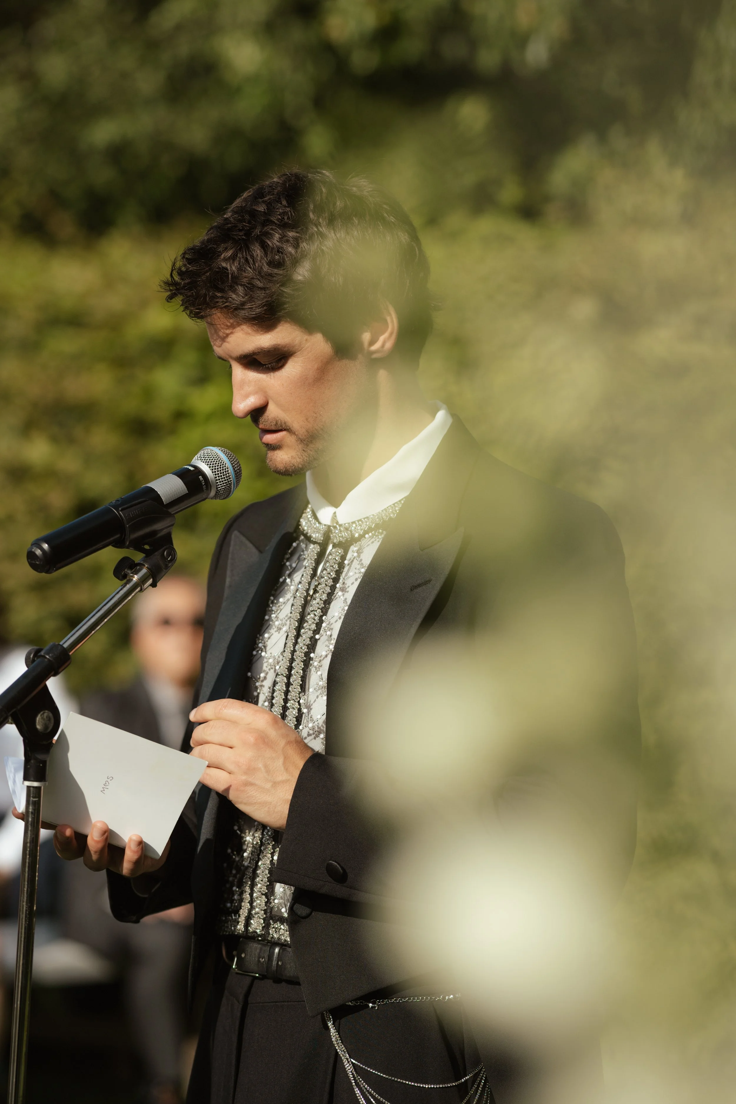 A man in a tuxedo standing outdoors, reading from a paper into a microphone during a formal event, with blurred greenery in the background.