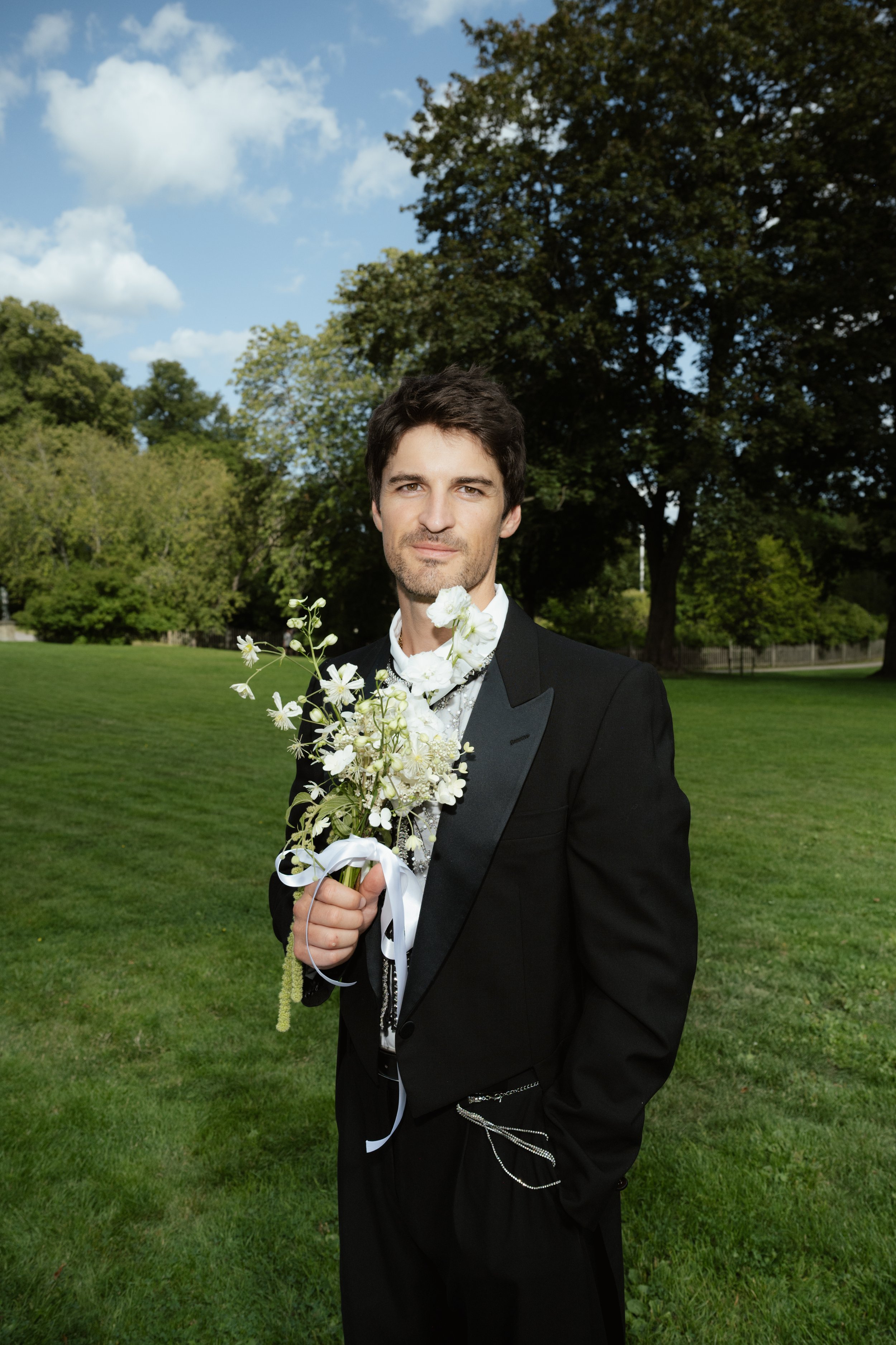 Man in a tuxedo holding a bouquet of white flowers outdoors on a grassy field, with trees and a partly cloudy sky in the background.